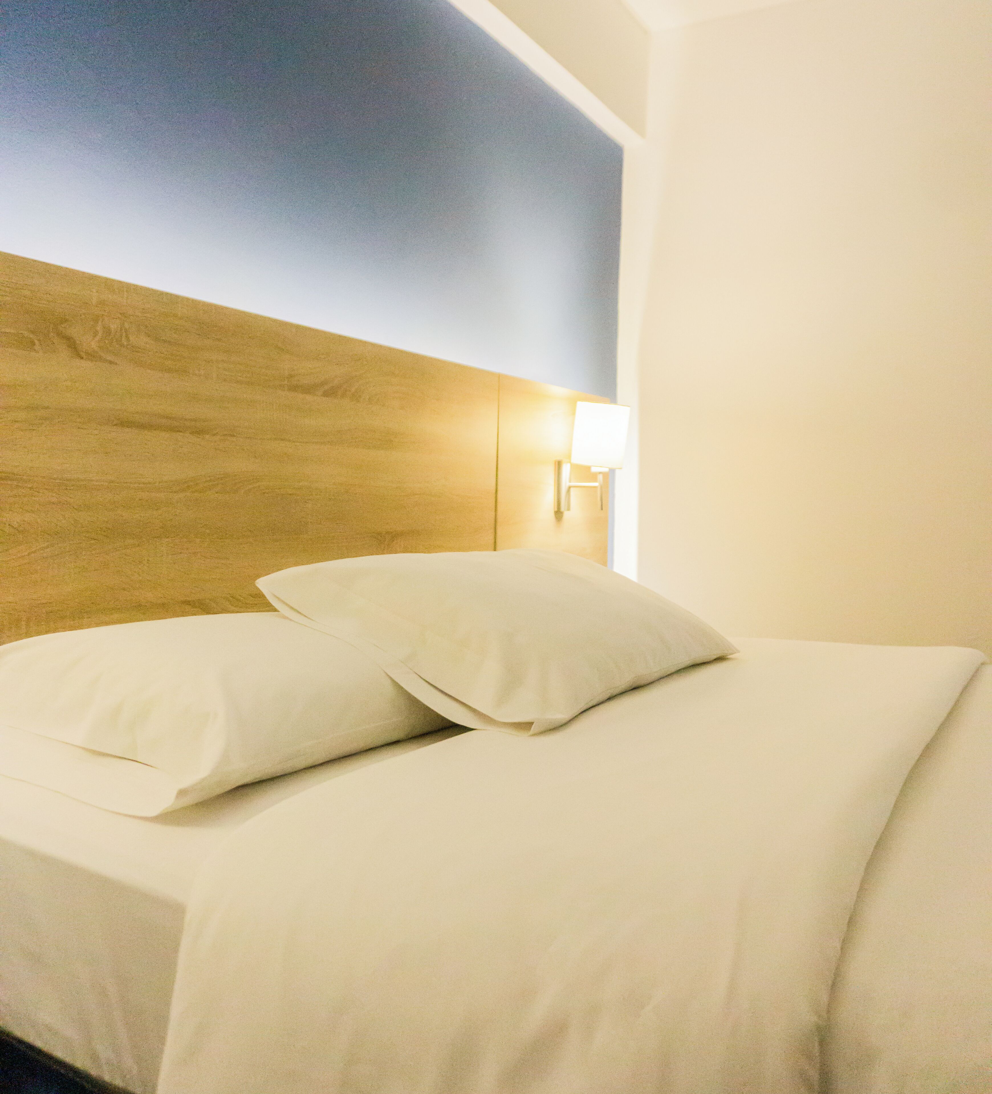 A neatly made bed with white linens and pillows, a wooden headboard, and a wall-mounted light fixture.