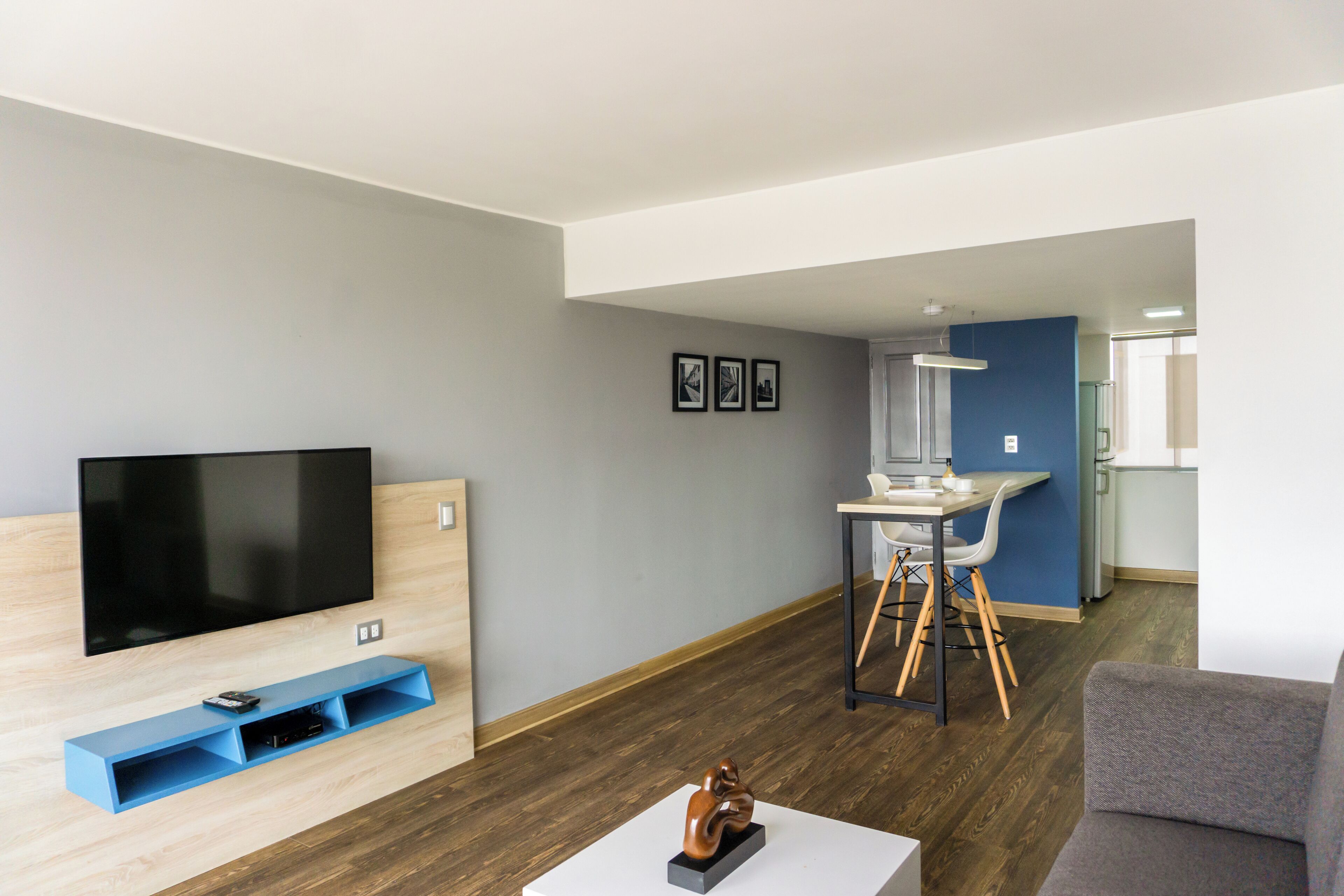 A modern living room with a flat-screen TV, a blue entertainment unit, a gray sofa, and a dining area with a white table and chairs.