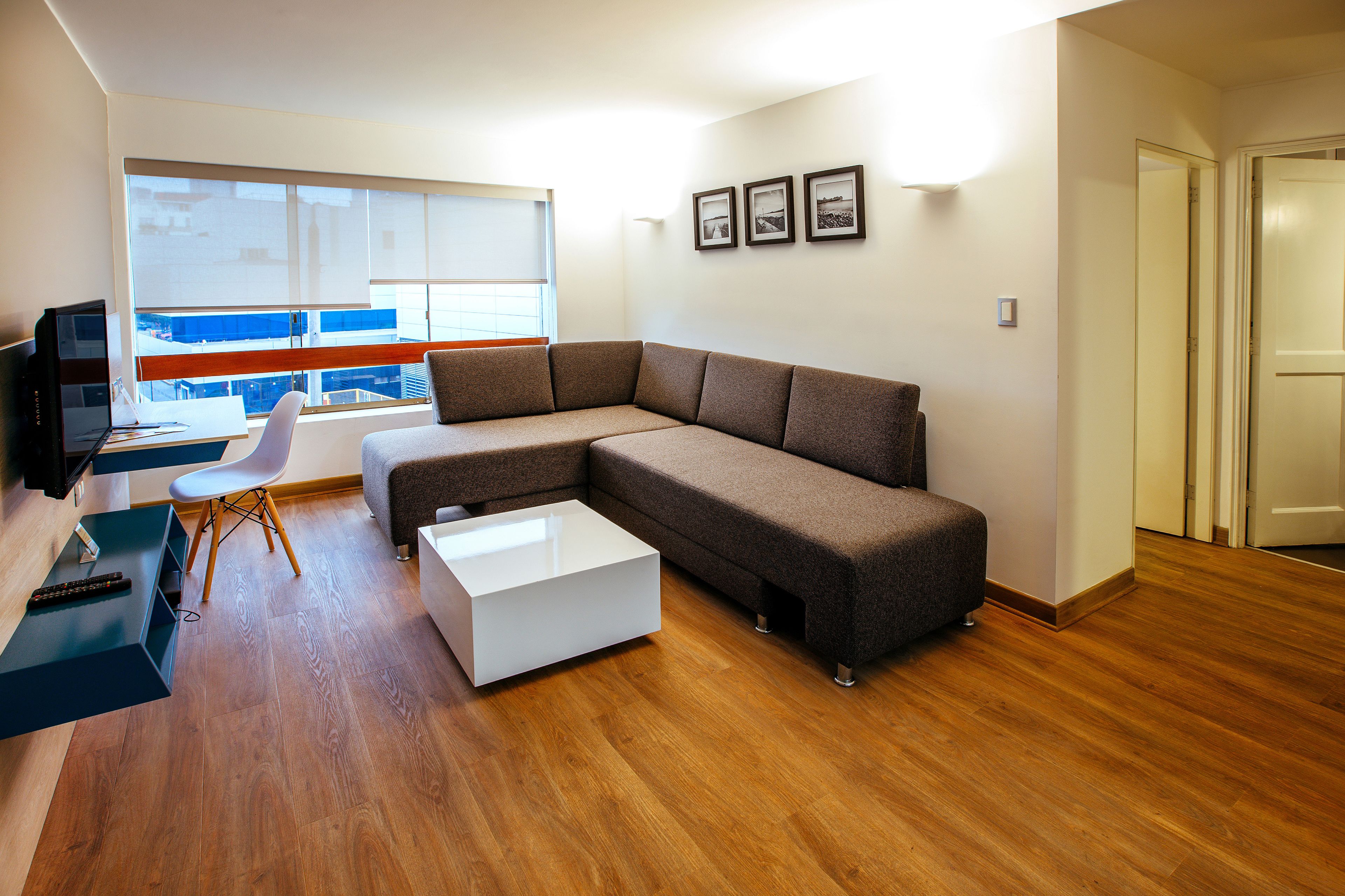 A modern living room with a sectional sofa, a white coffee table, a TV, and a large window with blinds.