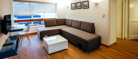 A modern living room with a sectional sofa, a white coffee table, a TV, and a large window with blinds.