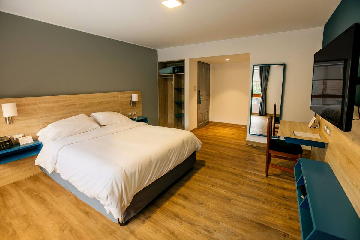 A modern hotel room with a large bed, bedside tables, a desk with a chair, a mirror, and a TV.