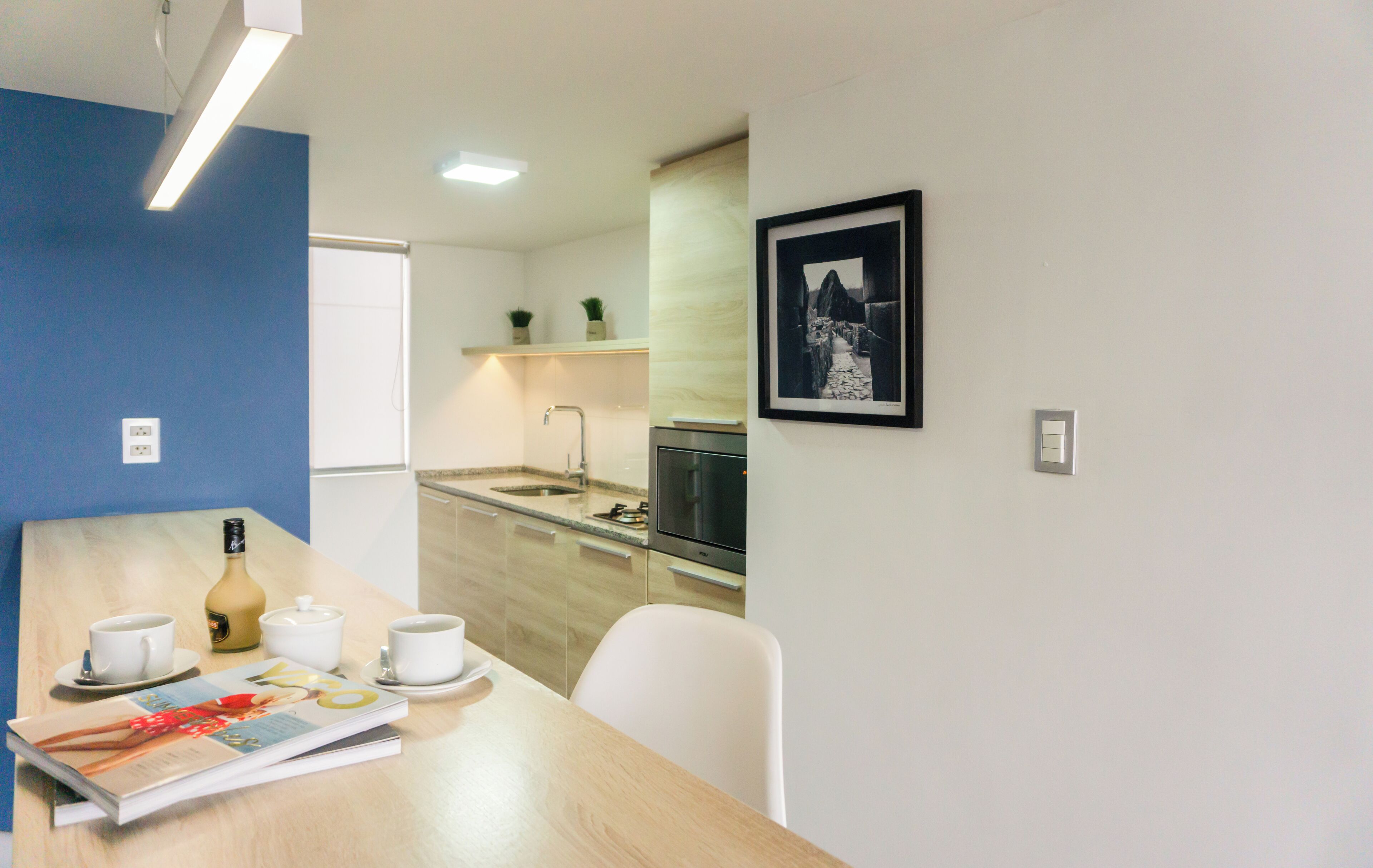 A modern kitchen with a sink, stove, and microwave. There is a dining table with cups and a magazine.