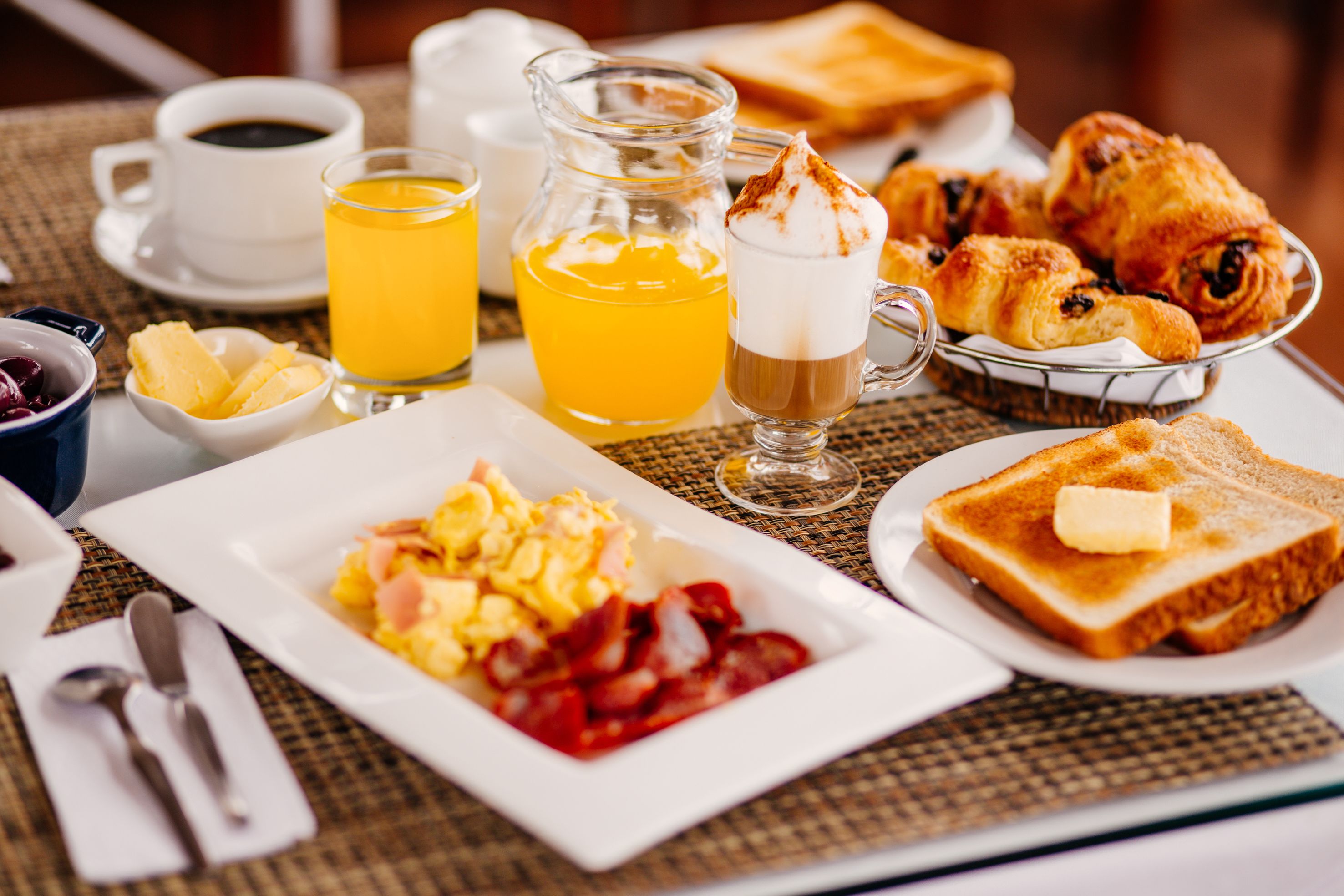 Una mesa de desayuno con huevos revueltos, tostadas, croissants y una jarra de jugo de naranja.