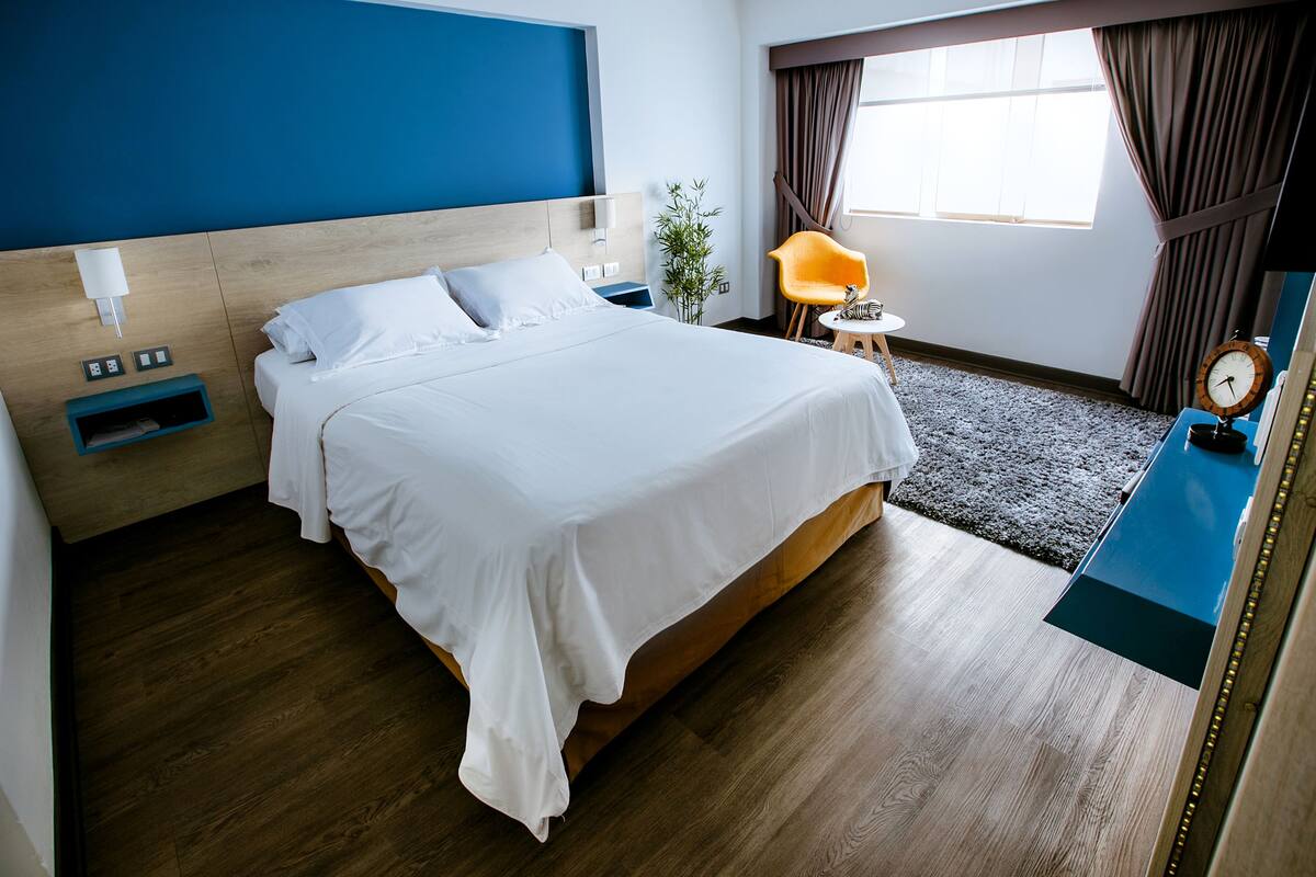 A hotel room with a large bed, a bedside table with a lamp, a small table with a clock, and a view of the outdoors through a window.