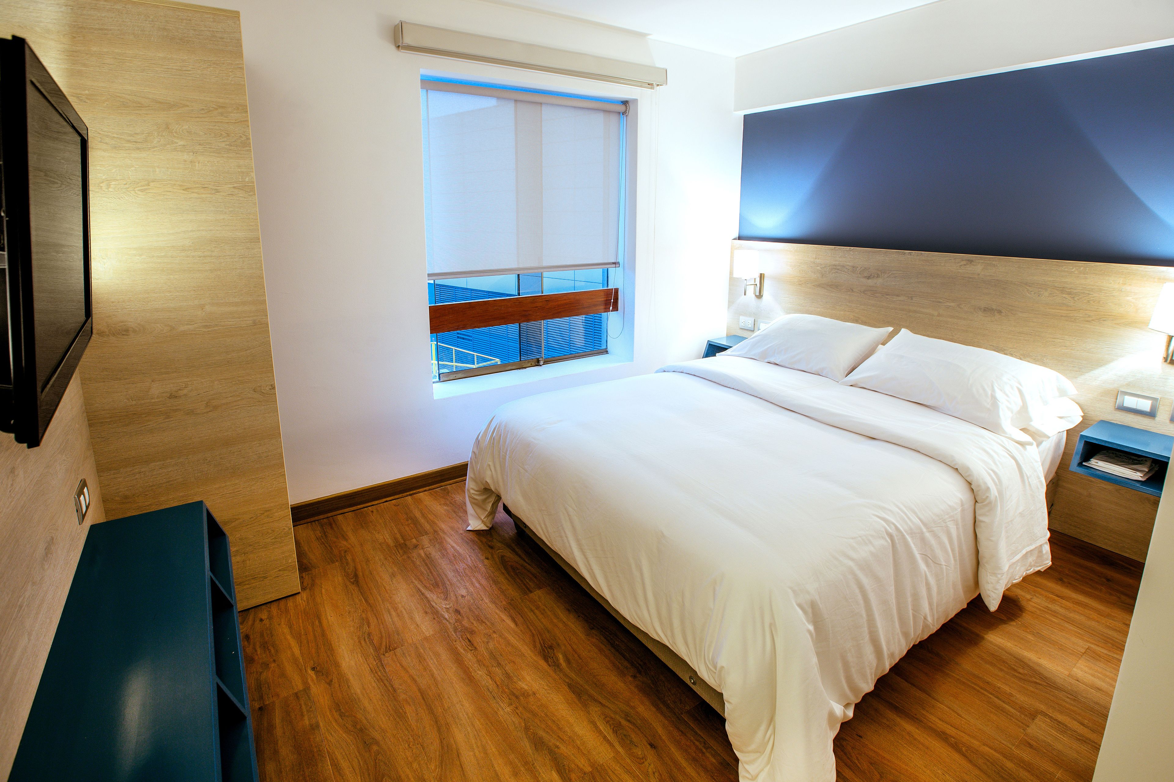 A hotel room with a wooden floor, a large bed with white bedding, a nightstand with a lamp, a TV mounted on the wall, and a window with blinds.