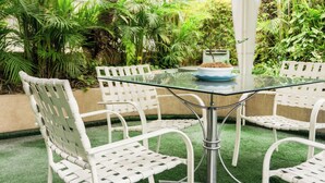 A glass-top table surrounded by white wicker chairs in an outdoor garden area with lush green plants and a palm tree. - Daniel's Apart Hotel (Lima)