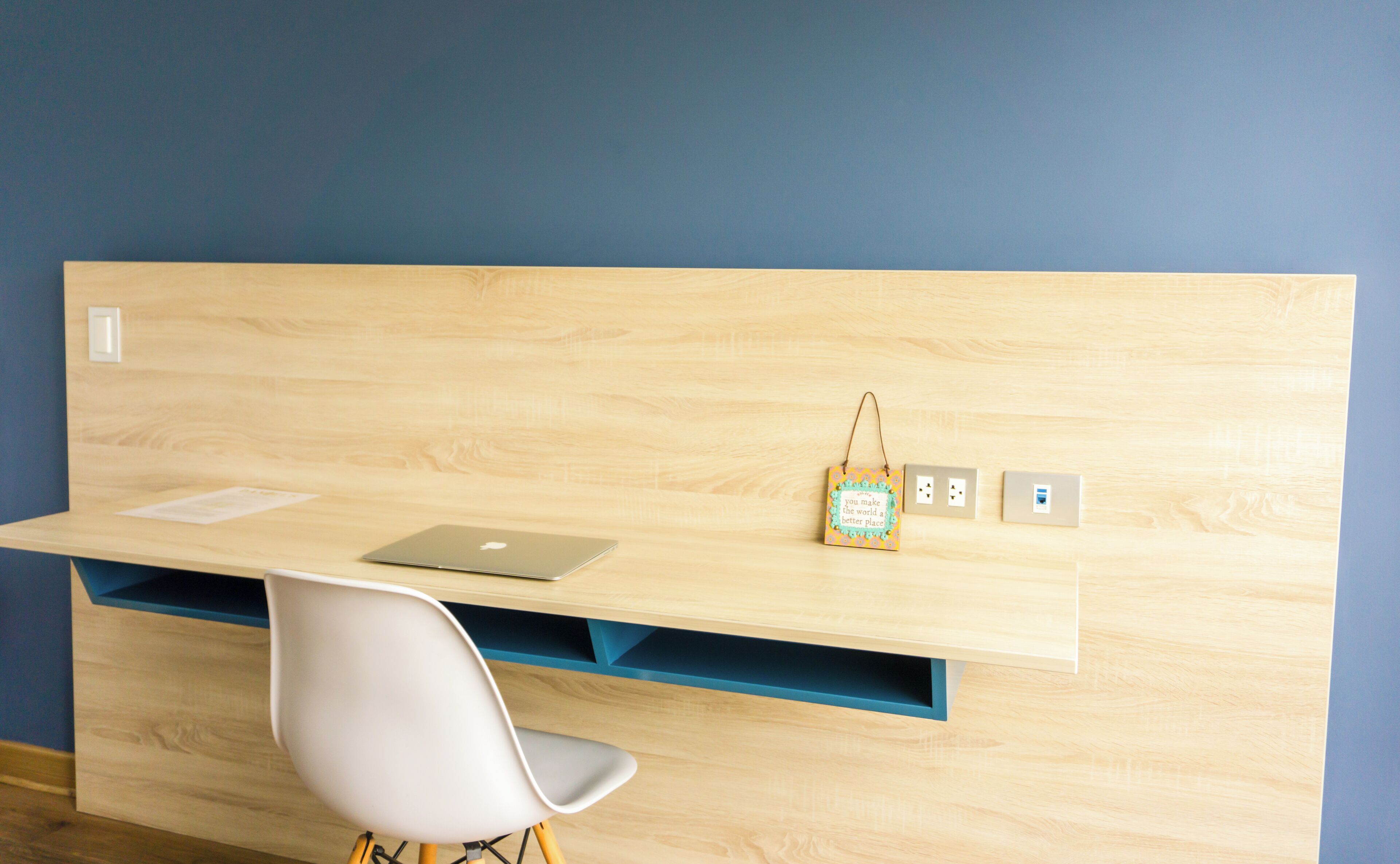 Un espacio de trabajo moderno con un escritorio de madera, una computadora portátil, una silla blanca y una frase motivacional en la pared.