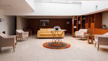 A spacious lobby with a yellow sofa, a round table with a vase, and a wooden shelving unit with various items.