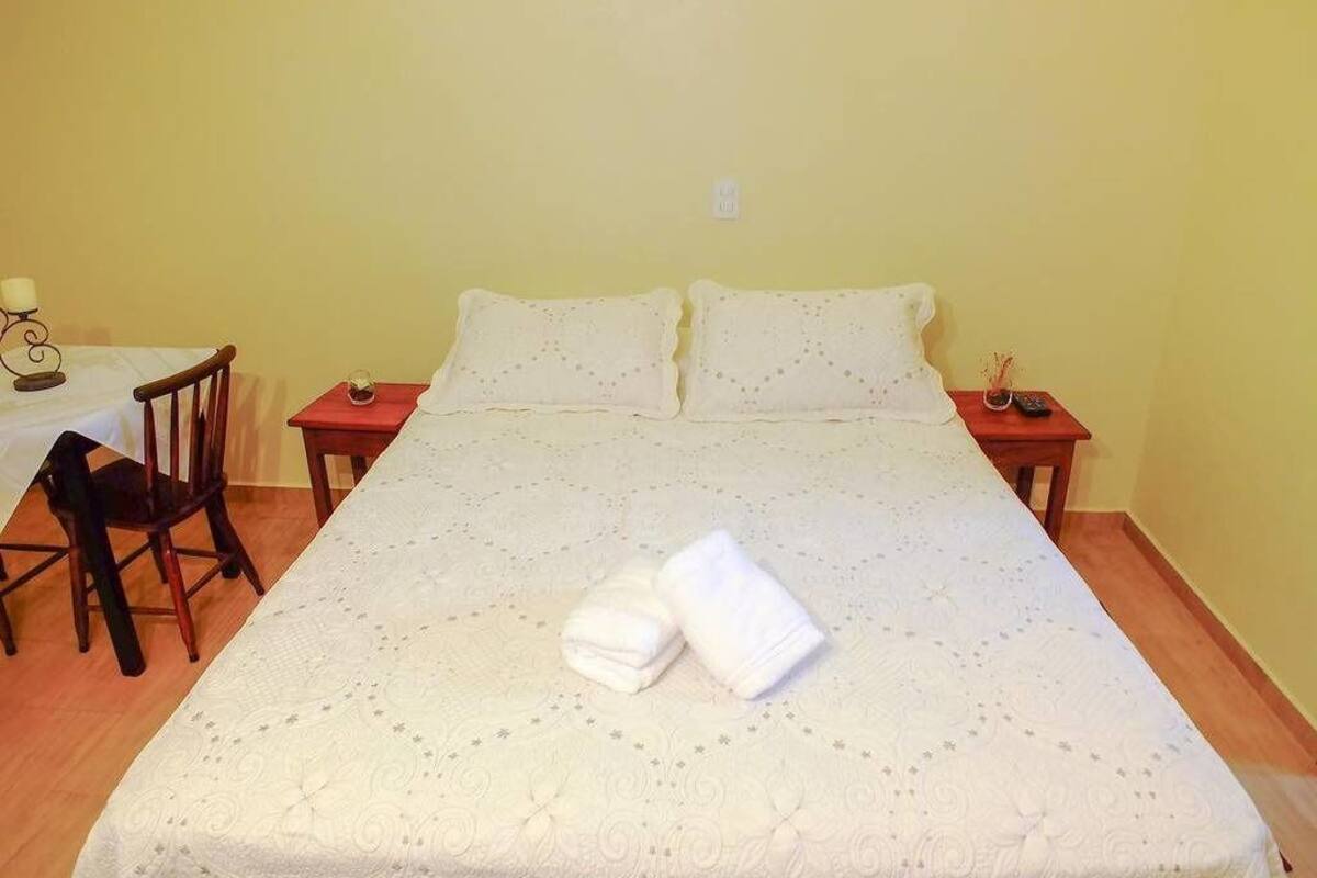 A neatly made bed with a white bedspread and two pillows, a wooden chair, a small table with a candle, and a nightstand with a lamp.