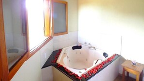 A bathroom with a large bathtub, a window, and a small table with a glass container. - Pousada Vista da Serra (Camanducaia)