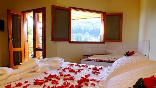 A hotel room with a bed, a window with a view of trees, a bathtub, and a door. - Pousada Vista da Serra (Camanducaia)