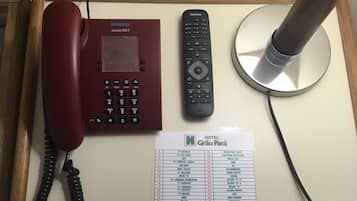 A hotel reception desk with a red telephone, a black remote control, and a cable management device. A hotel reception sign lists TV channels and their corresponding channel numbers.