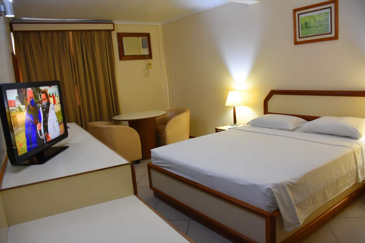 Habitación de hotel con cama, televisor en un escritorio, silla, mesita de noche y lámpara.