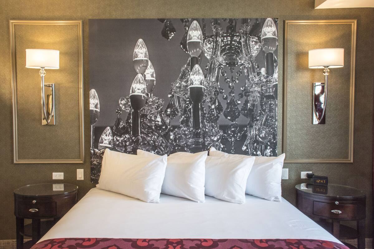 A hotel room with a large bed, white pillows, a red patterned bedspread, two bedside tables, a nightstand, a wall clock, and a large abstract painting above the bed.
