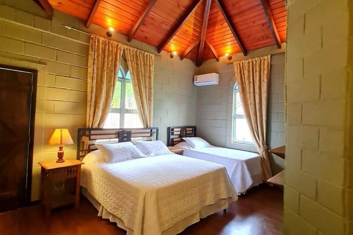 A room with two beds, wooden ceiling beams, a window with curtains, a wall-mounted air conditioner, and a door.