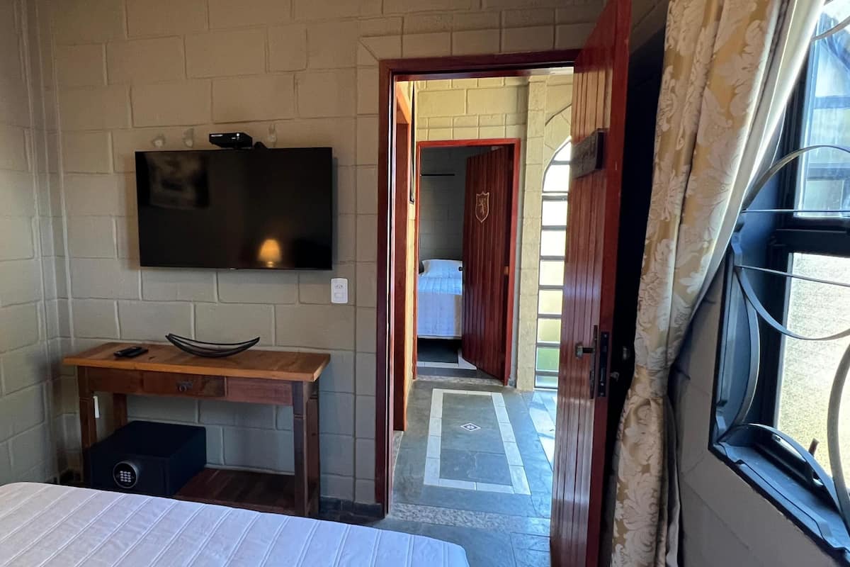 A bedroom with a bed, a TV mounted on the wall, a desk with a computer, and a door leading to another room.