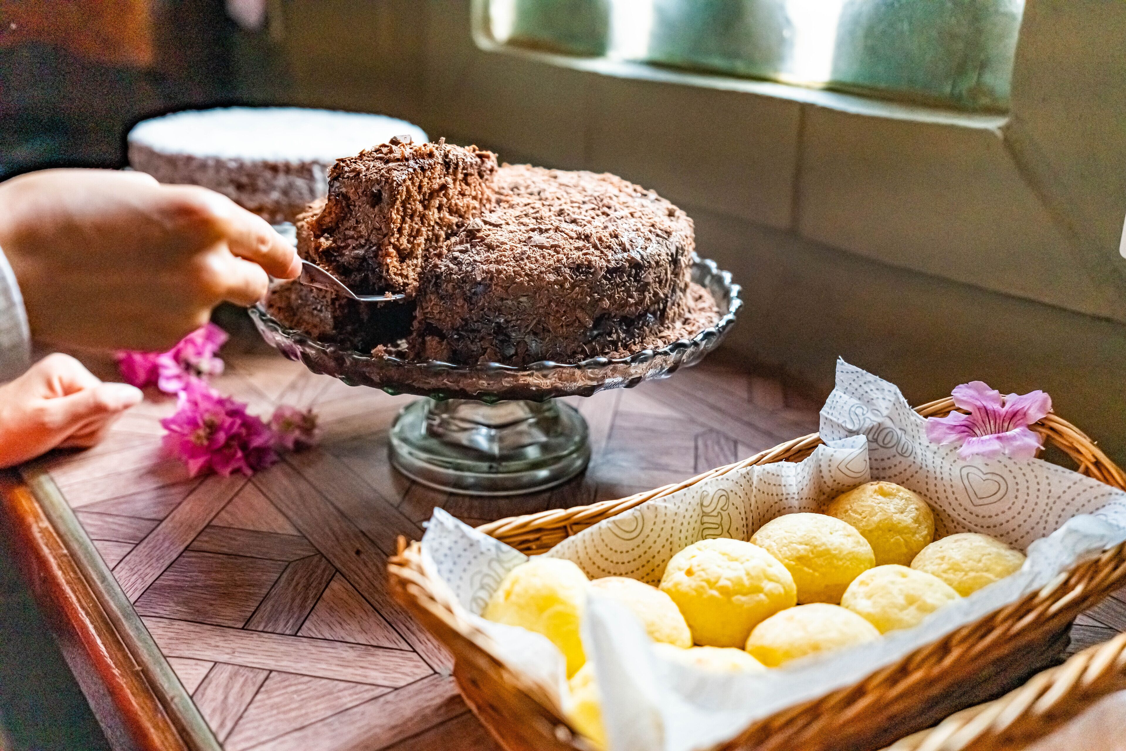 Taart op een glazen etagère, een hand reikt ernaar en een mand met gele donuts staat op een houten tafel.