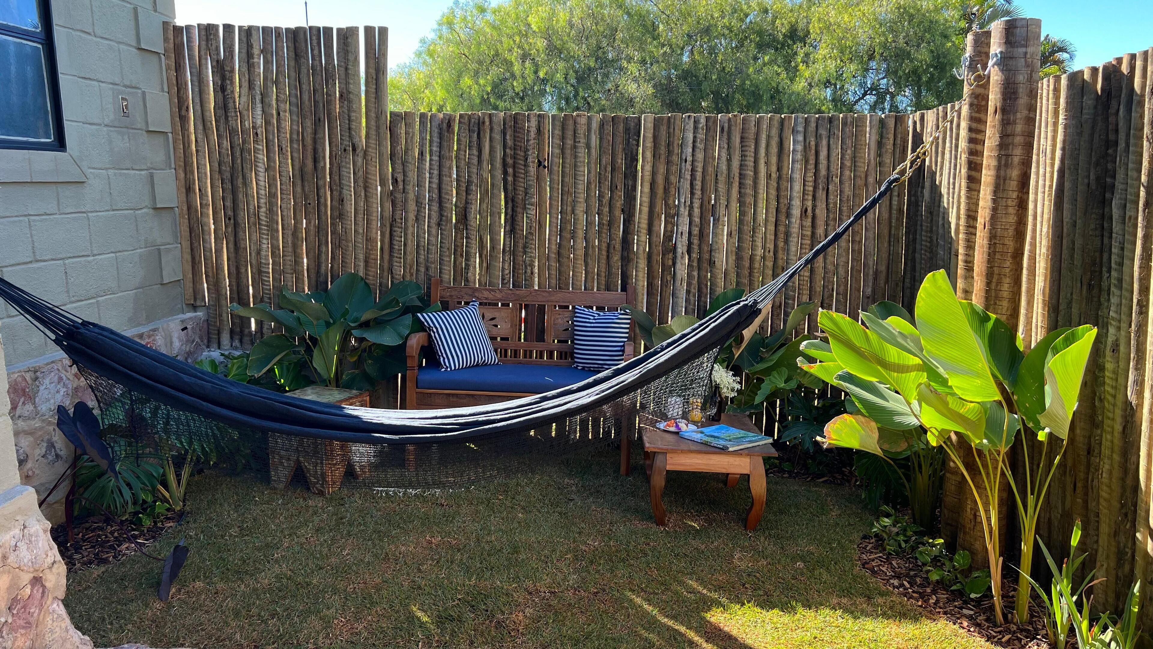 A hammock set up between two wooden posts in a backyard with a wooden fence, a wooden bench, and various plants.