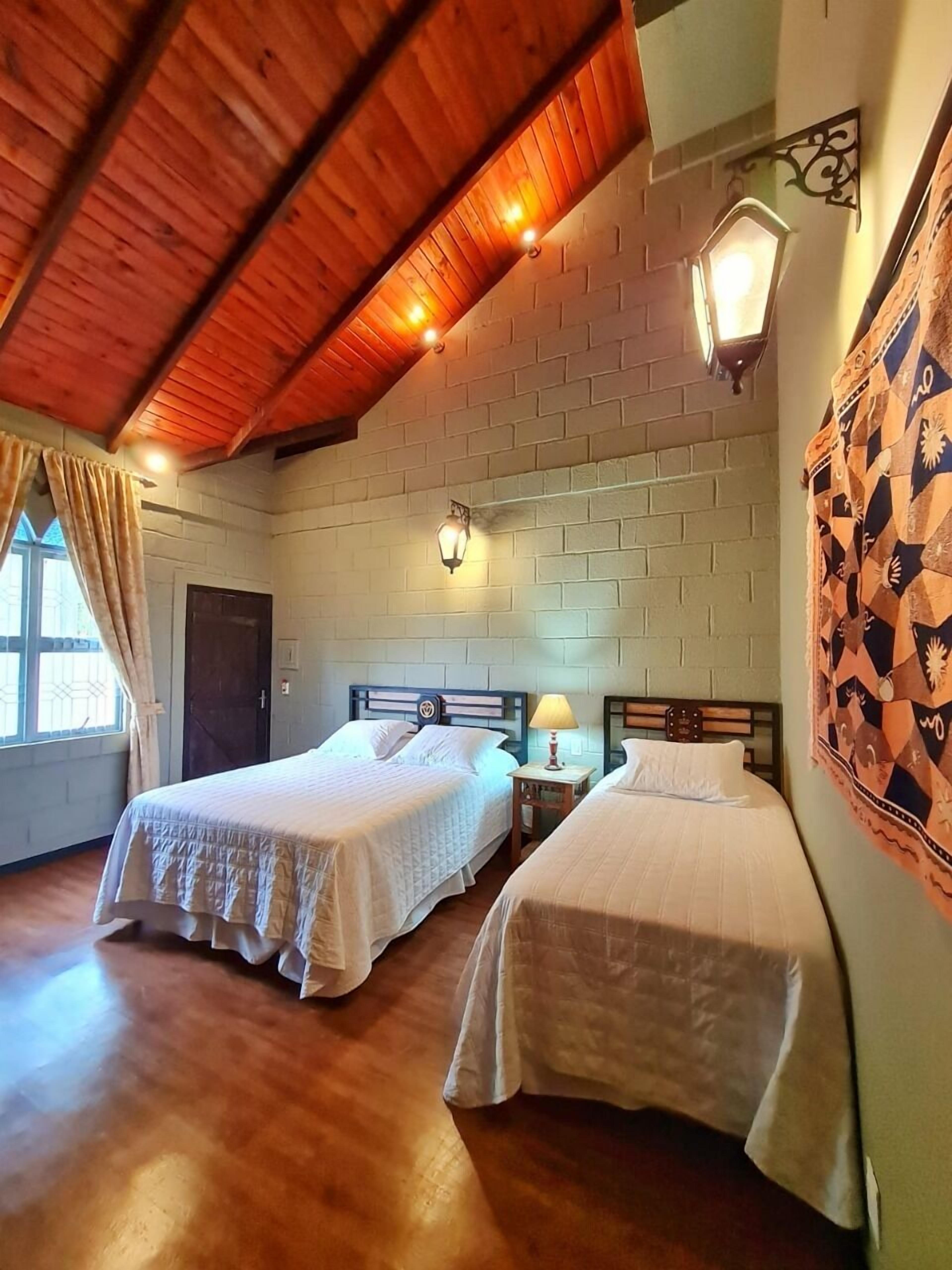A room with two single beds, a brick wall, wooden ceiling beams, and a window with curtains.