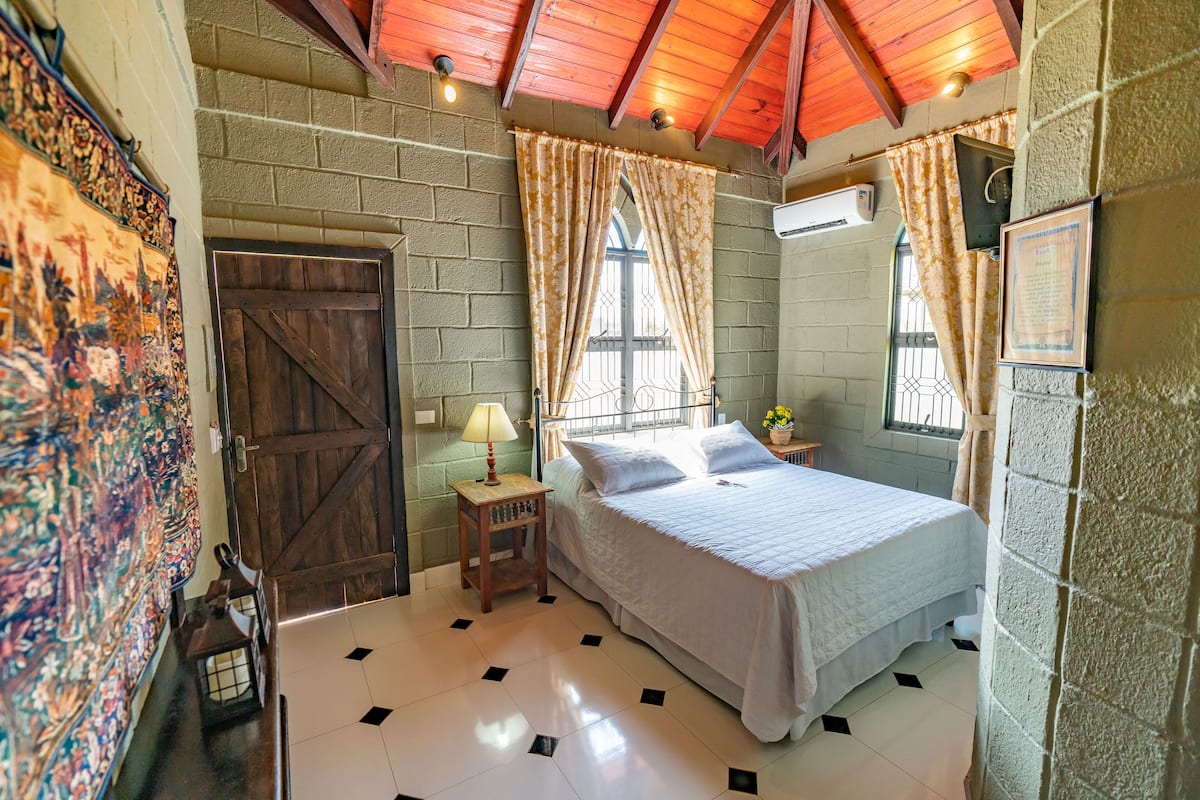 A bedroom with a wooden door, a bed with white bedding, a nightstand with a lamp, and a window with curtains.