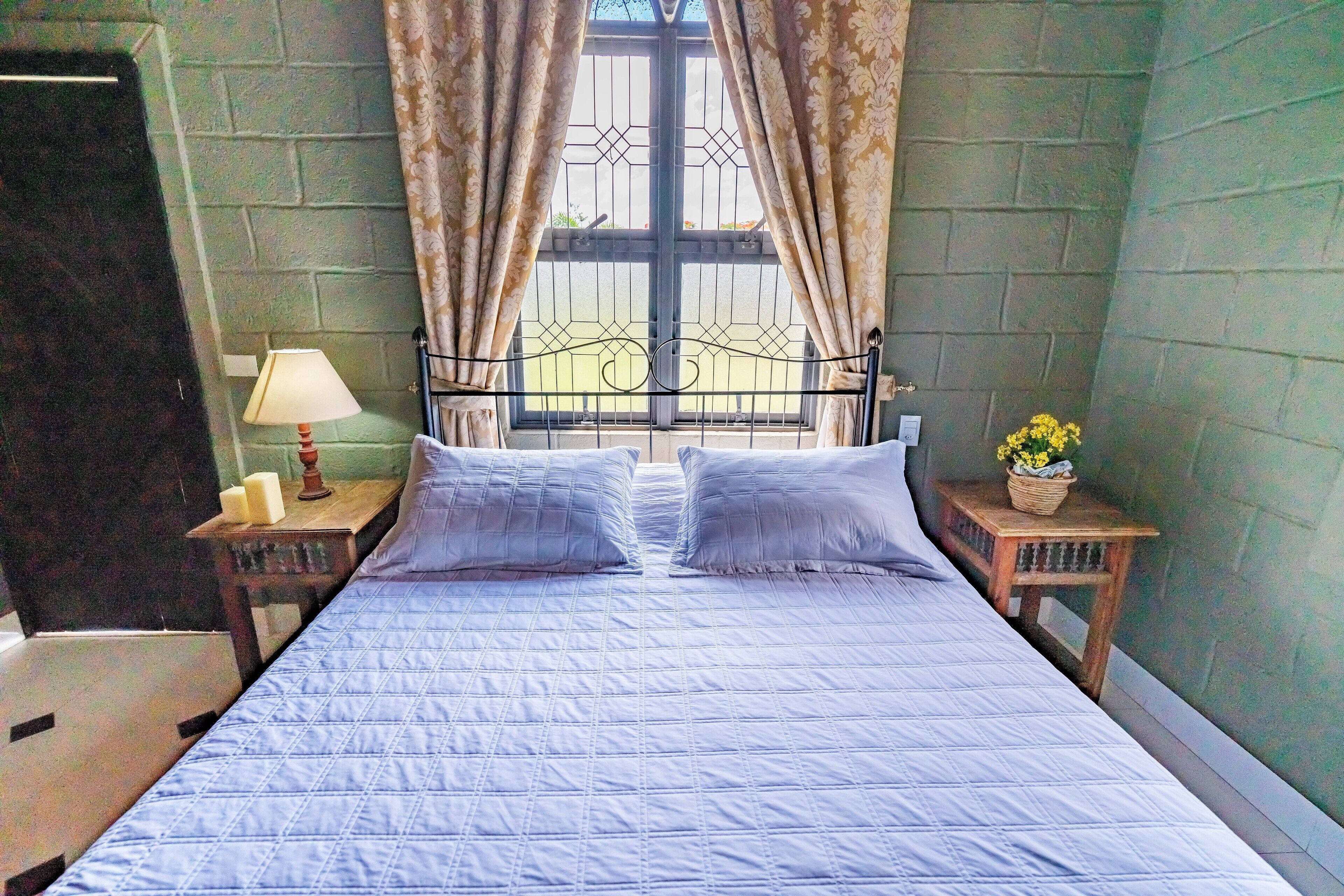 A bed with a quilted blue cover, two pillows, a nightstand with a lamp, and a window with curtains.