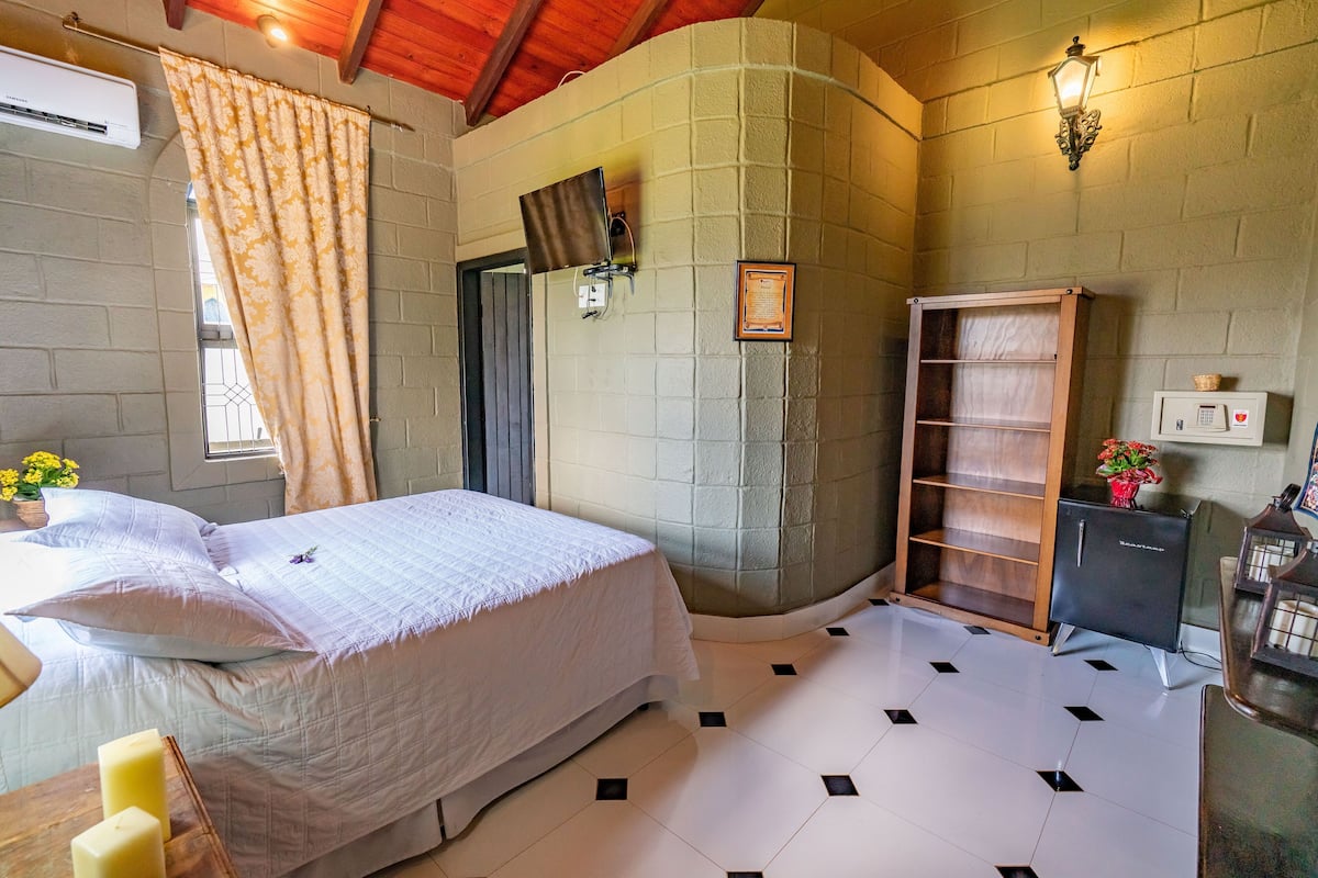 A bedroom with a bed, a bedside table with candles, a window with curtains, a wall-mounted TV, and a shelf with a plant.