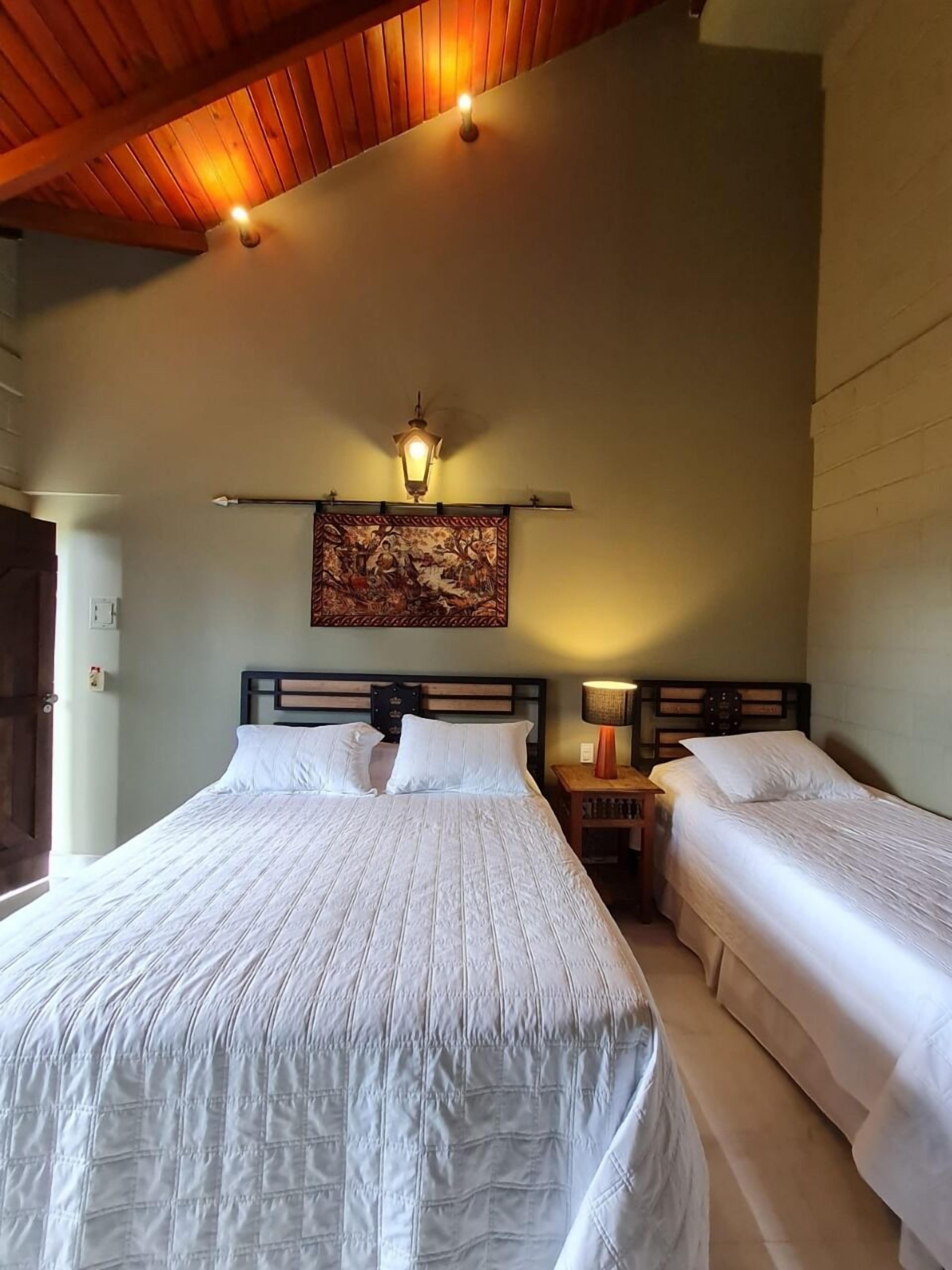 A bedroom with two beds, a wooden ceiling, a wall-mounted lamp, and a painting.