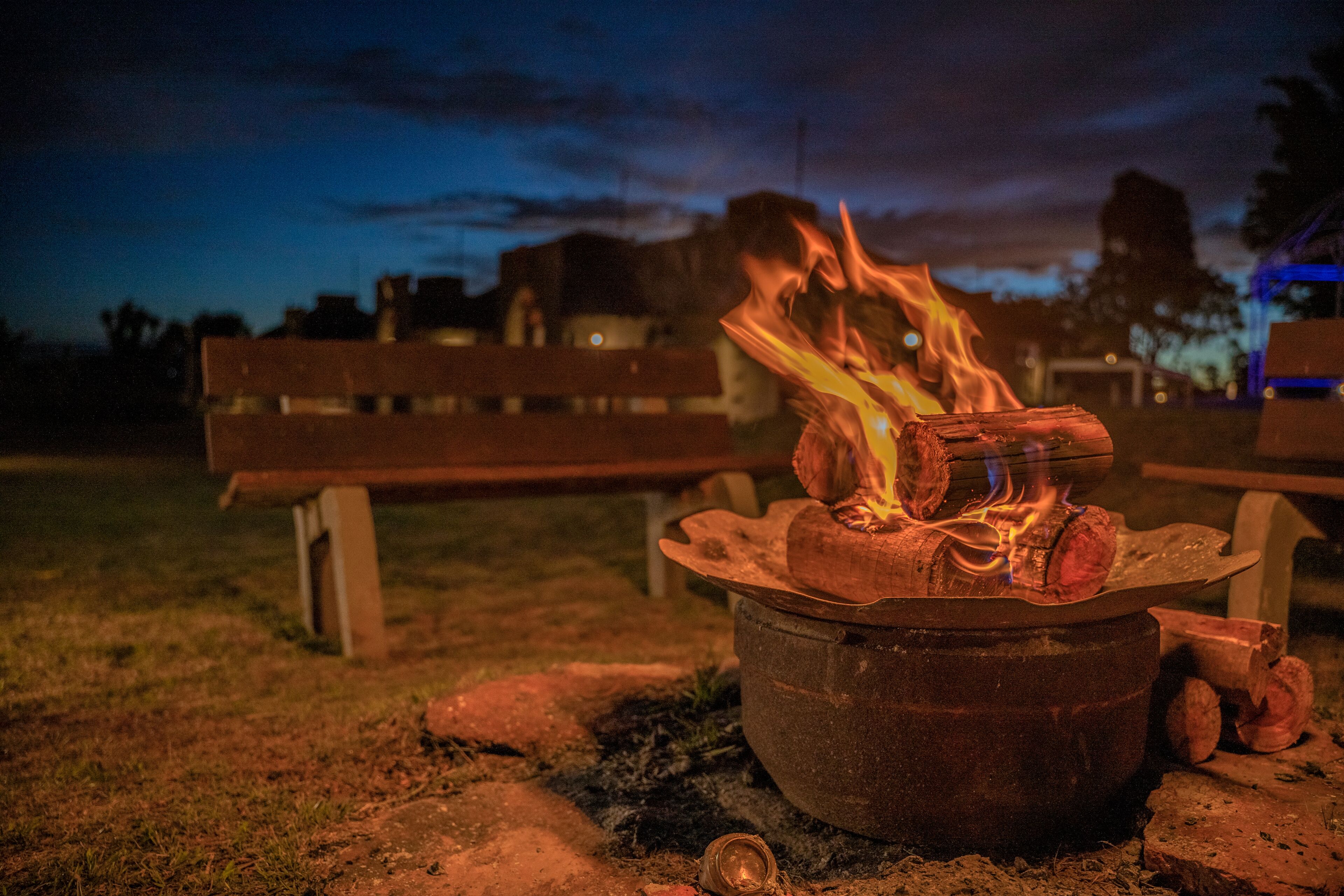 A campfire with logs and a pot, set on a grassy area with benches in the background.