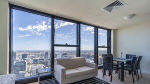 A modern living room with a large window, a sofa, a dining table with chairs, and a city view. - StayIcon Serviced Apartment On Collins (Melbourne)