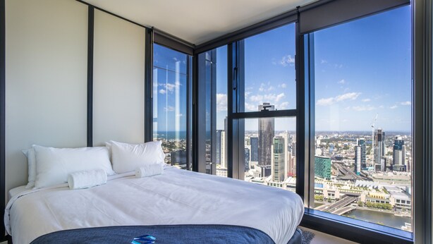 A modern hotel room with a large bed, white linens, and a view of a cityscape through floor-to-ceiling windows. - StayIcon Serviced Apartment On Collins (Melbourne)