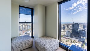 A corner of a room with a large window offering a city view, two ottomans with a patterned cover, and a ceiling light. - StayIcon Serviced Apartment On Collins (Melbourne)