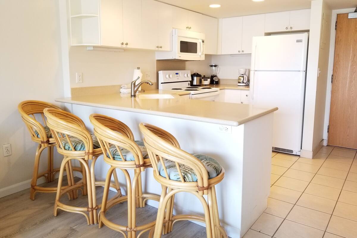 A kitchen with a central island, white cabinets, a microwave, a stove, and a refrigerator. There are three wicker chairs with cushions.