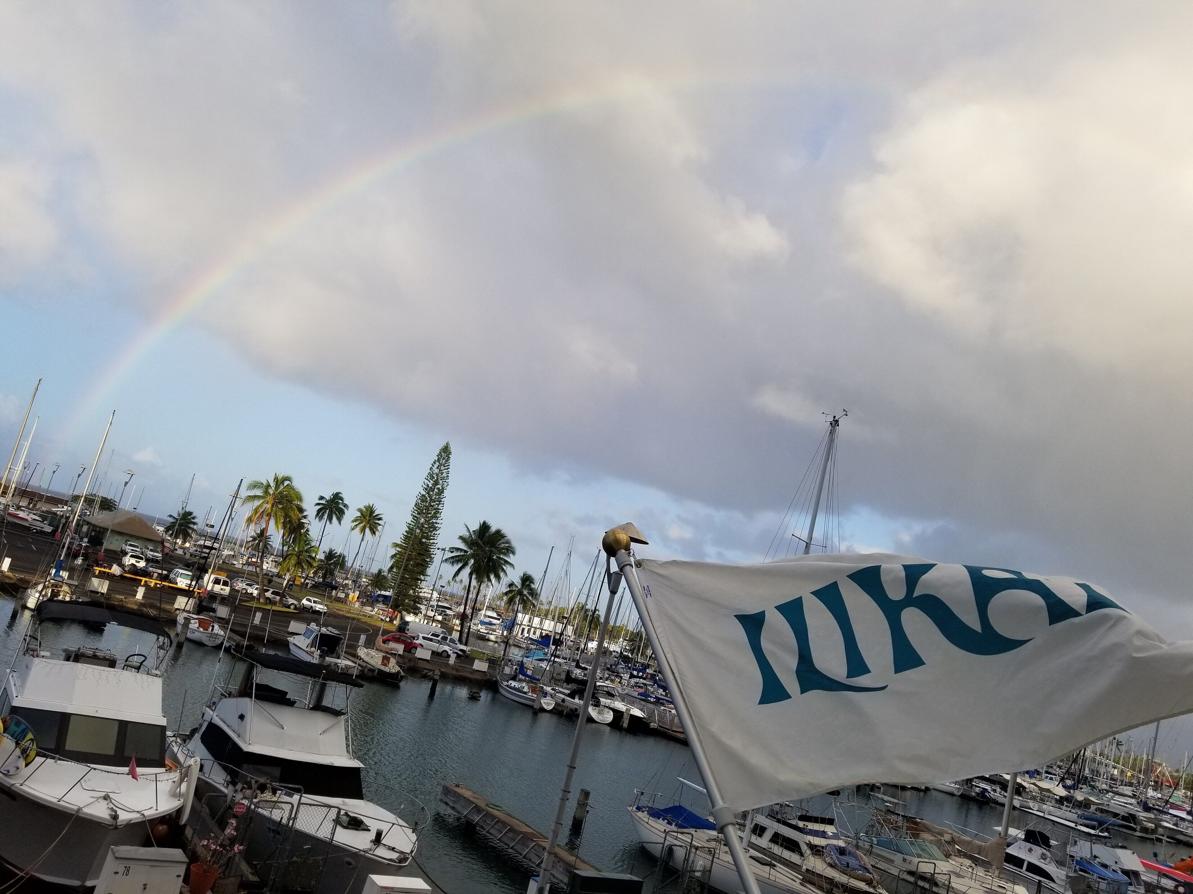 Un puerto deportivo con barcos amarrados, una bandera con un logotipo y un arcoíris en el cielo.