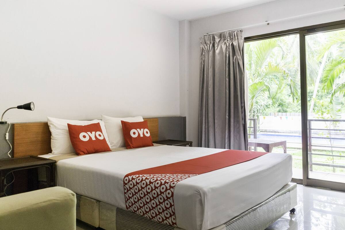A hotel room with a bed, a nightstand, a lamp, and a view of greenery through the window.