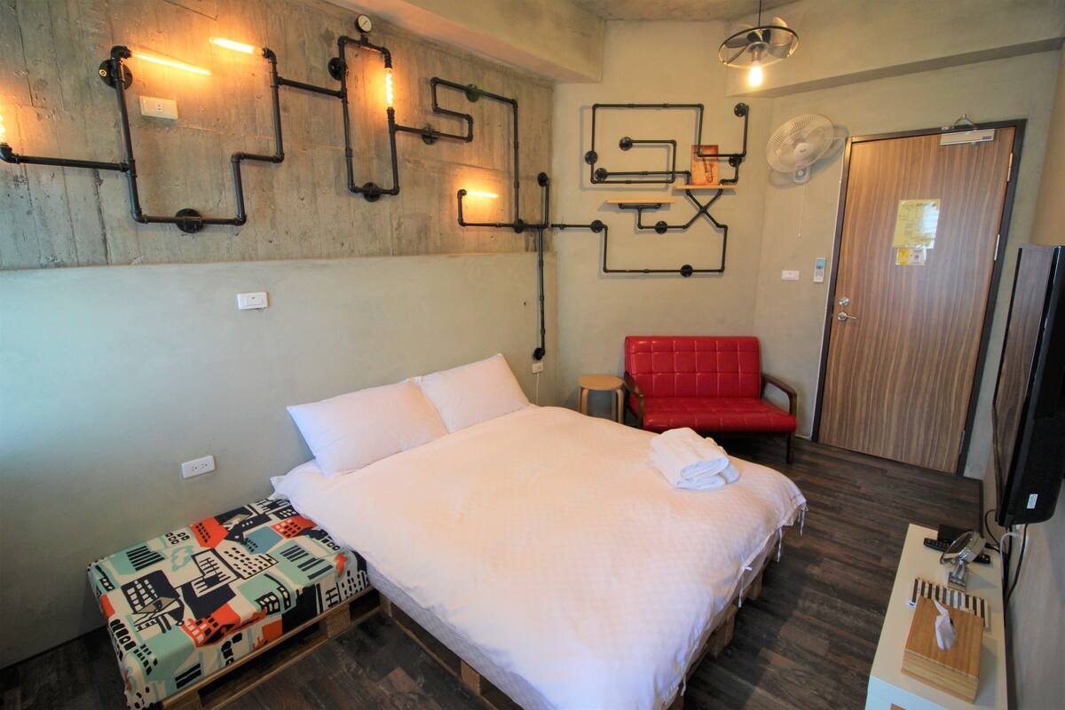 A hotel room with a bed, a red chair, a wooden bench, and a wall-mounted light fixture.