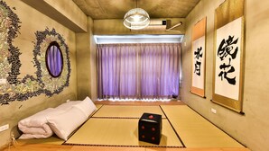 Une pièce avec un sol recouvert de tatamis, un futon servant de lit, un miroir décoratif, un luminaire suspendu et une vue sur l’extérieur par une fenêtre ornée de rideaux violets.
