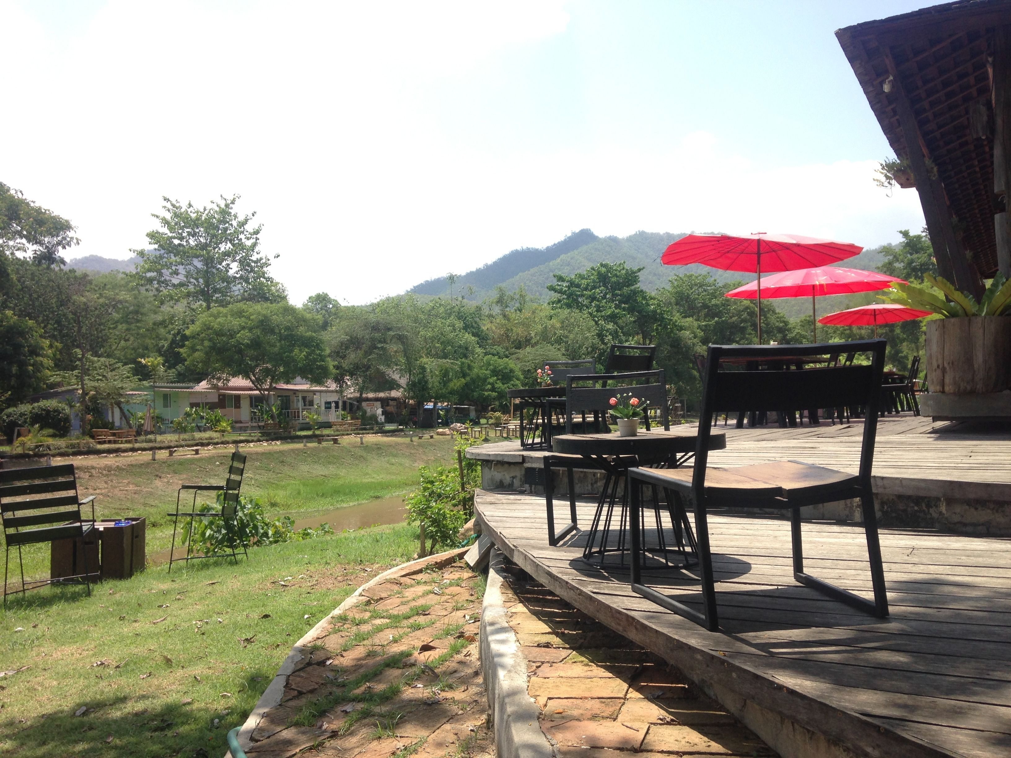 An outdoor seating area with wooden tables and chairs, surrounded by greenery and a view of hills.