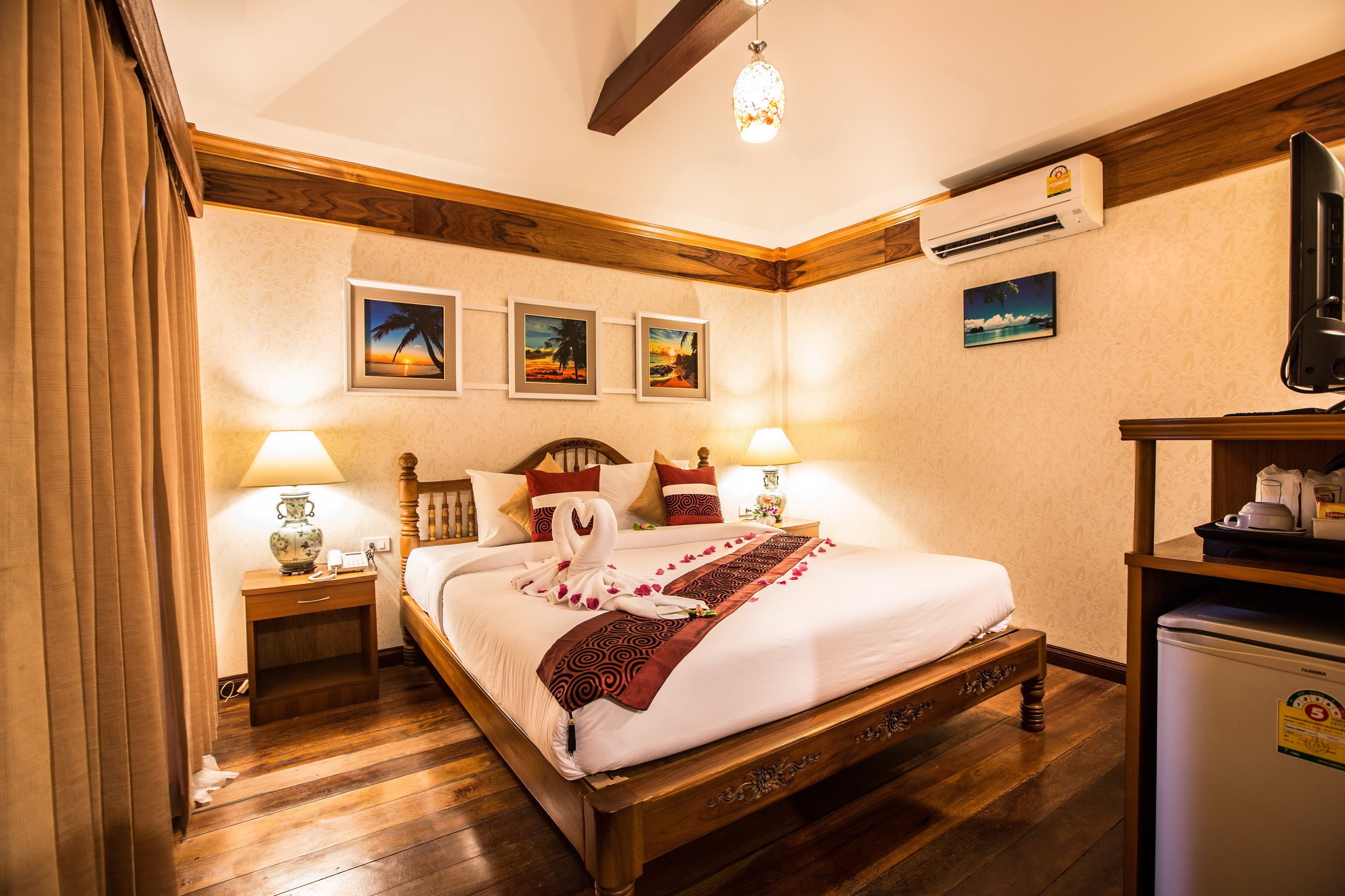 Una camera d'albergo con un letto in legno, due comodini con lampade, un condizionatore d'aria a parete e un frigorifero.