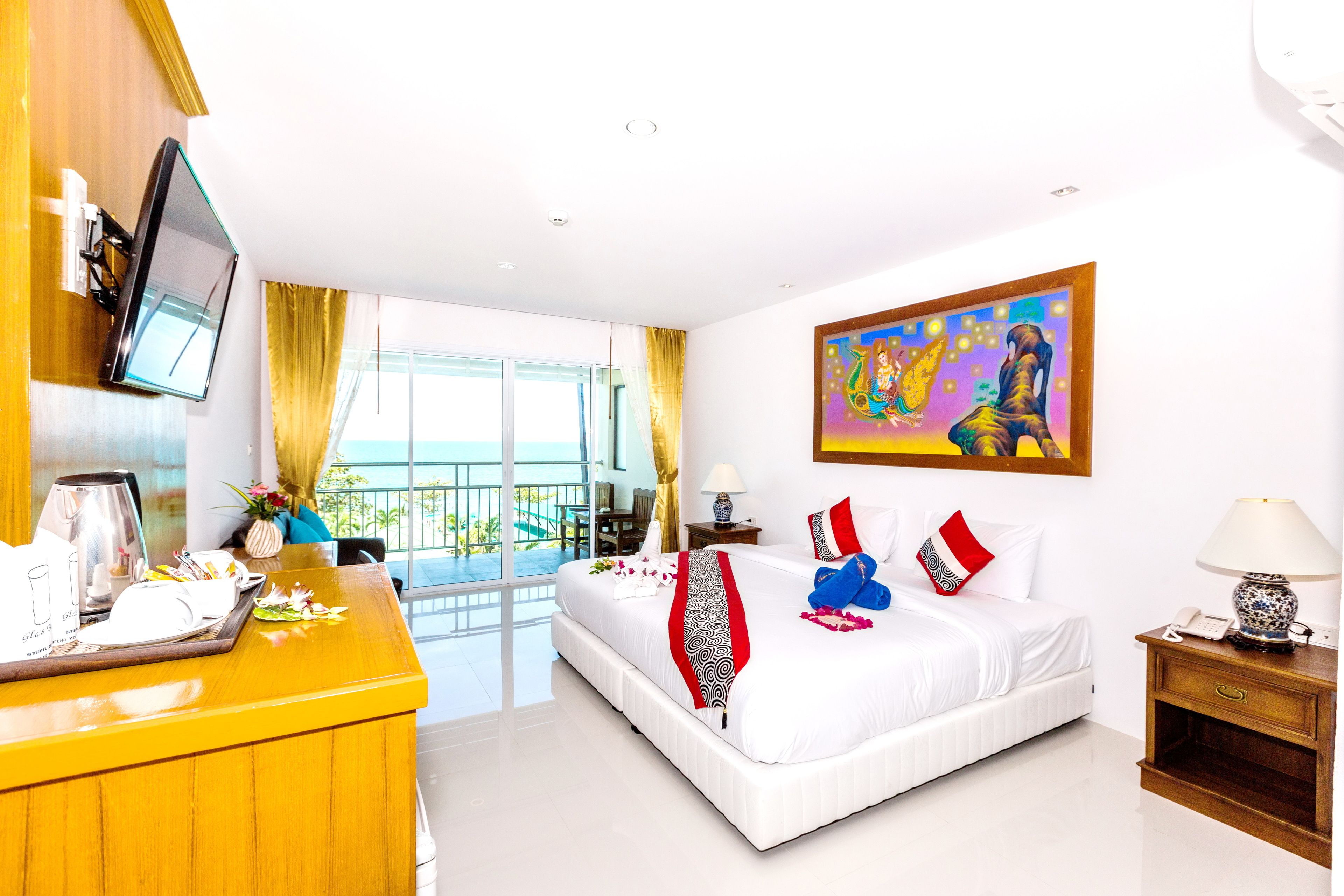 A hotel room with a bed, a desk with a flat-screen TV, a balcony with a view, and a painting on the wall.
