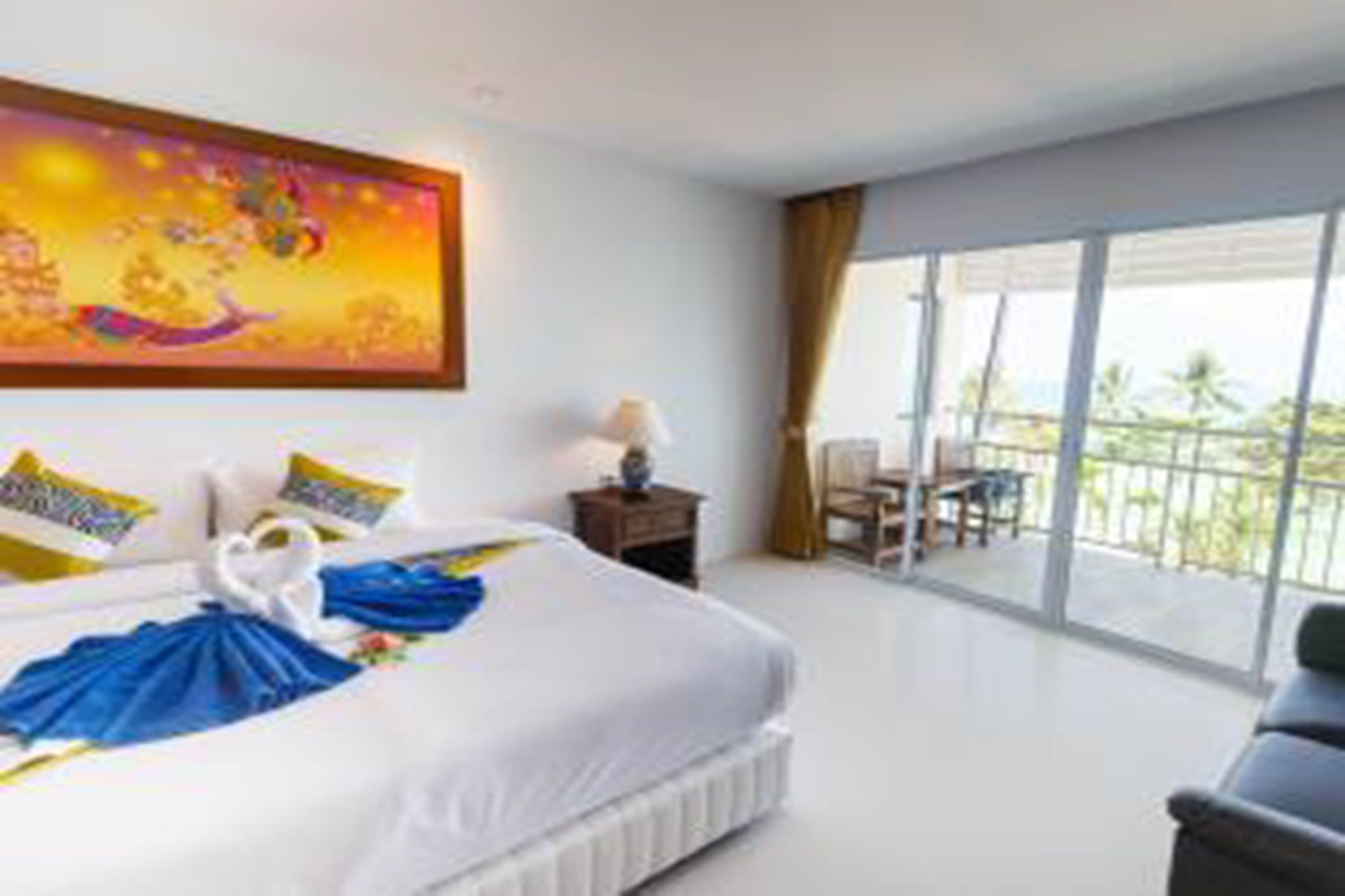 A hotel room with a large bed, a painting on the wall, a balcony with a view, and a desk with a lamp.