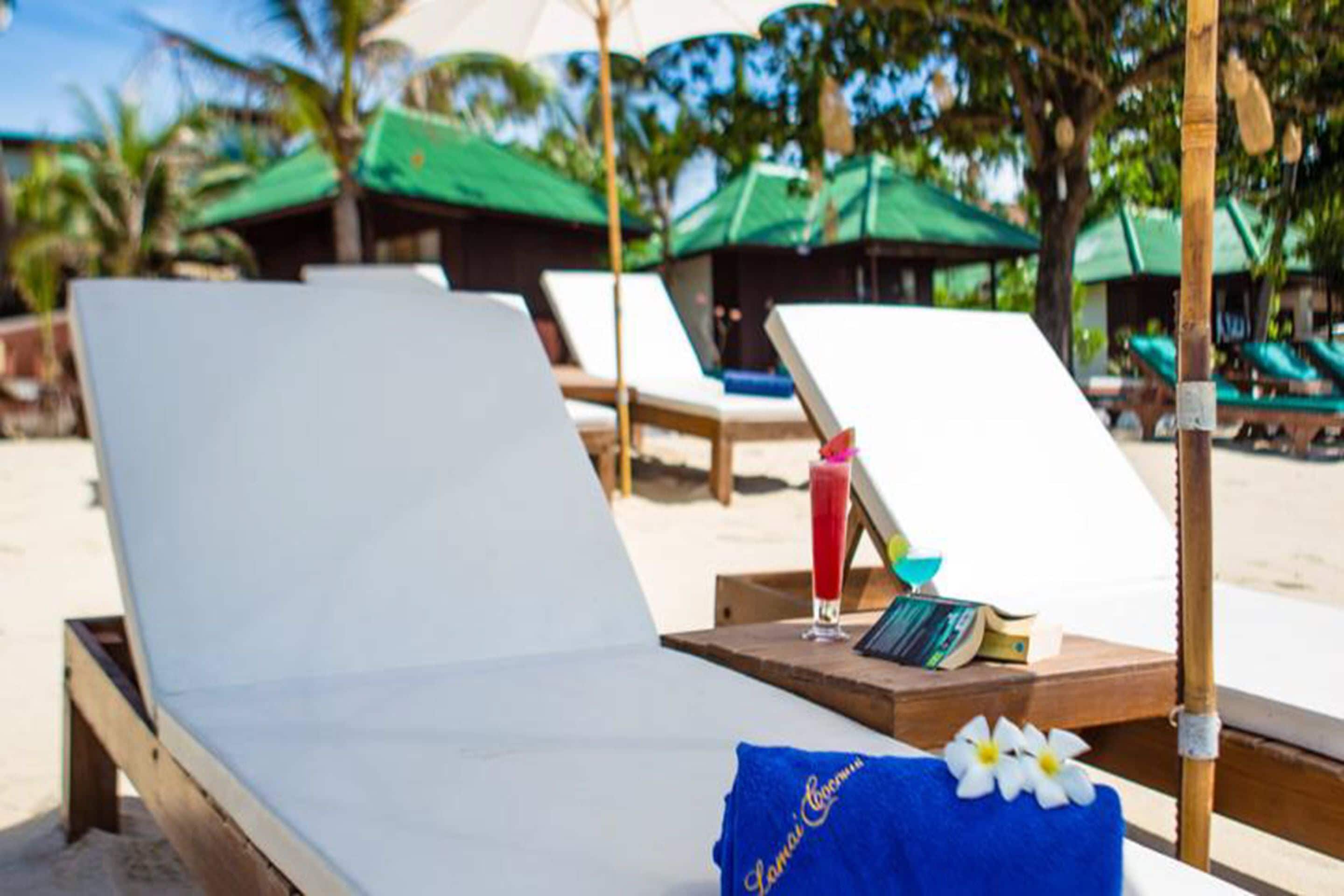 Habitación de hotel frente a la playa, con una cama blanca, una toalla azul y una bebida tropical sobre la mesita.