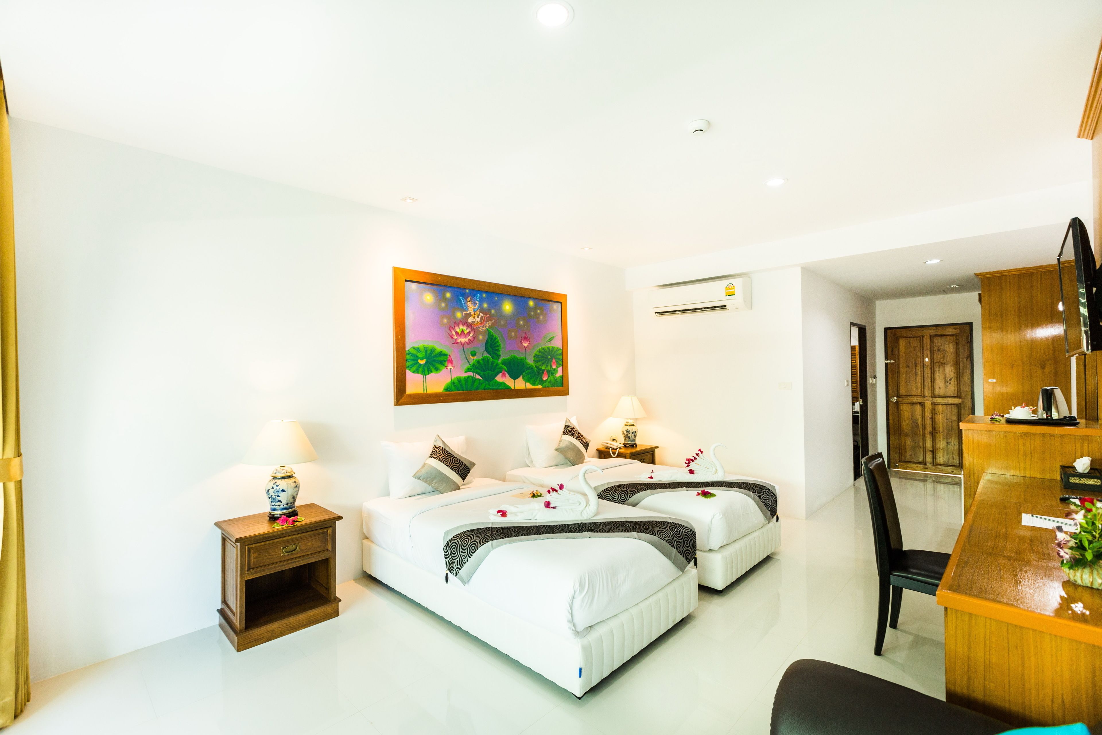 A hotel room with two beds, a desk, a chair, a small table, a painting, and an air conditioning unit.