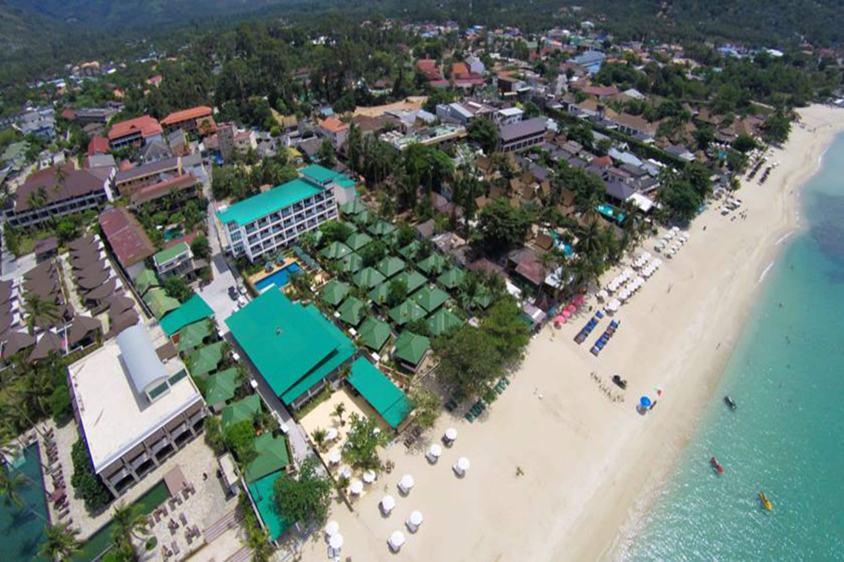 An aerial view of a coastal area with a beach, buildings, and a hotel with a green roof.