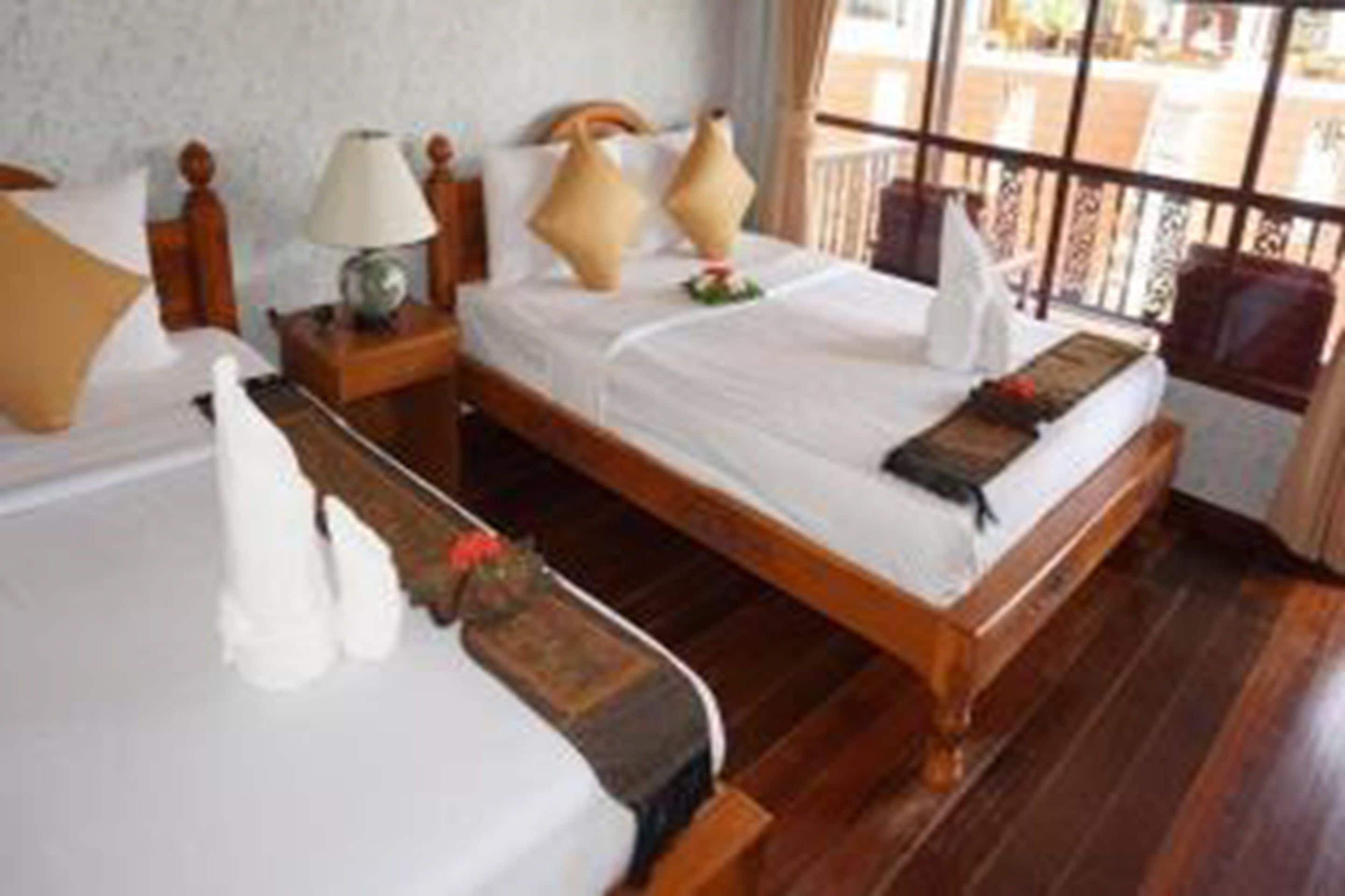 A hotel room with a wooden bed, white bedding, a nightstand with a lamp, and a view of the outdoors through large windows.