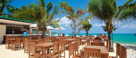 An outdoor seating area with wooden tables and chairs, surrounded by palm trees and a view of the beach.