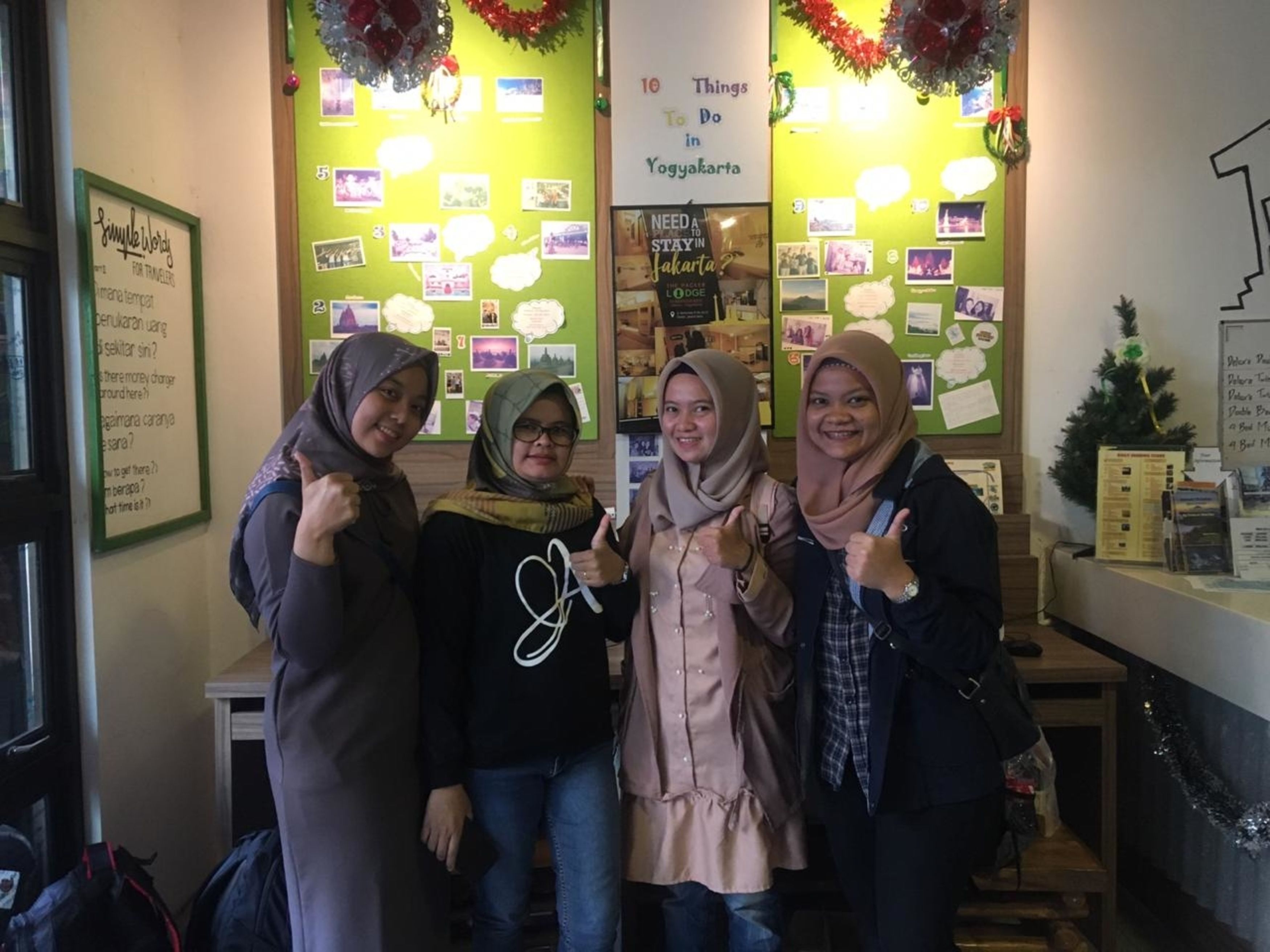 A reception area with a green wall displaying a '10 Things To Do in Yogyakarta' sign, a small Christmas tree, and a bulletin board with photos and notes.