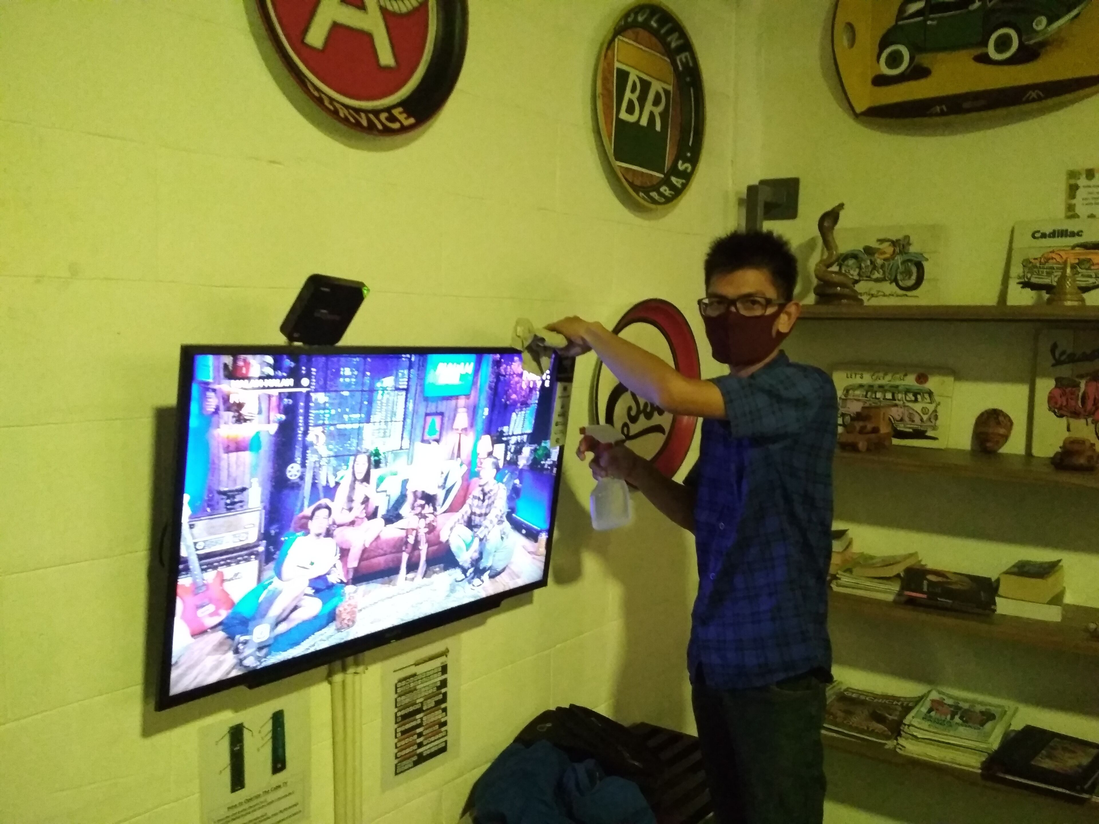 A room with a television mounted on the wall displaying a show. There are shelves with various items including books and figurines.