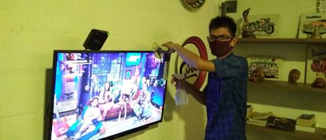 A room with a television mounted on the wall displaying a show. There are shelves with various items including books and figurines.