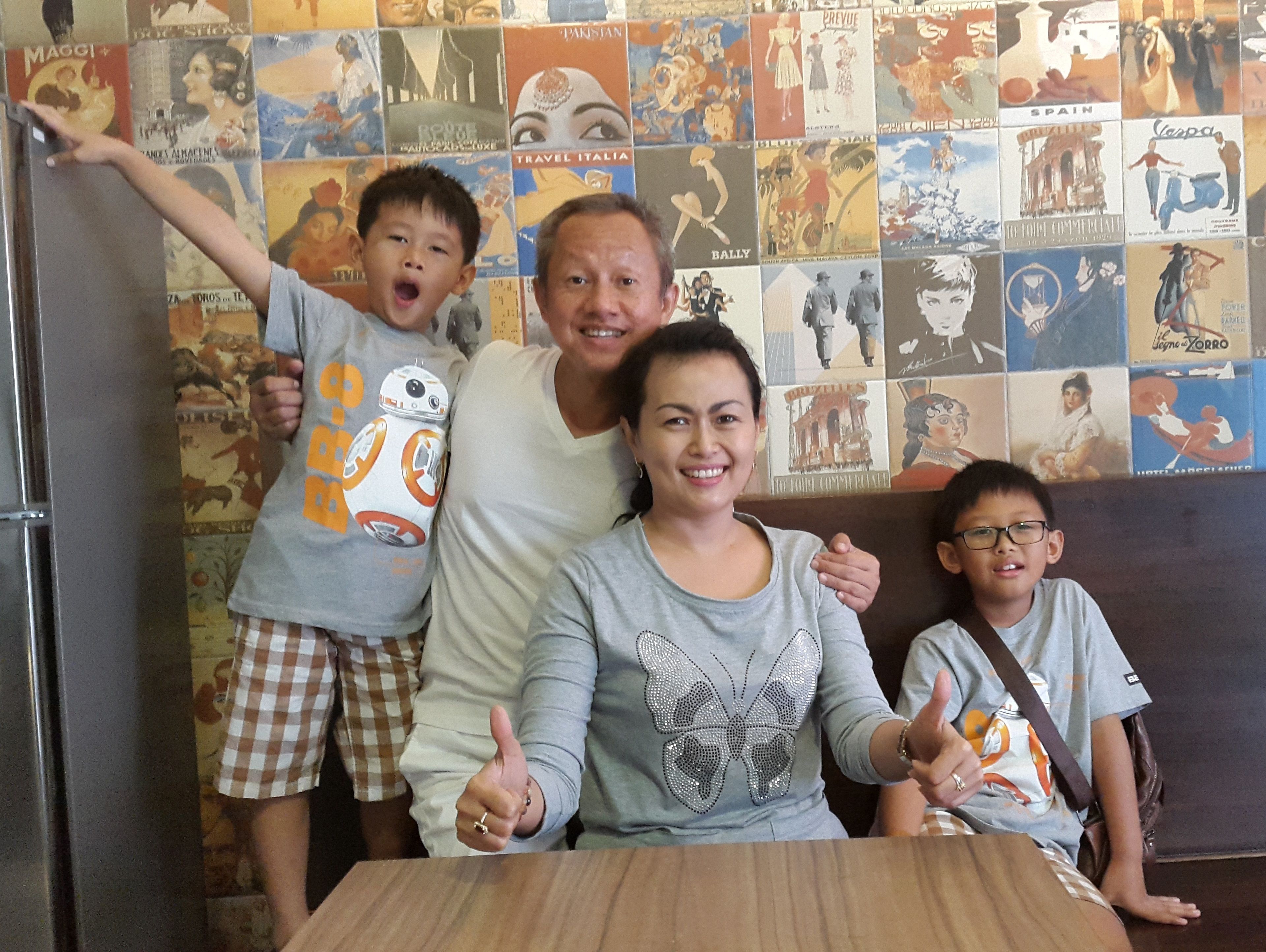 Une famille est assise à une table, avec un mur recouvert d’affiches de voyage en arrière-plan.