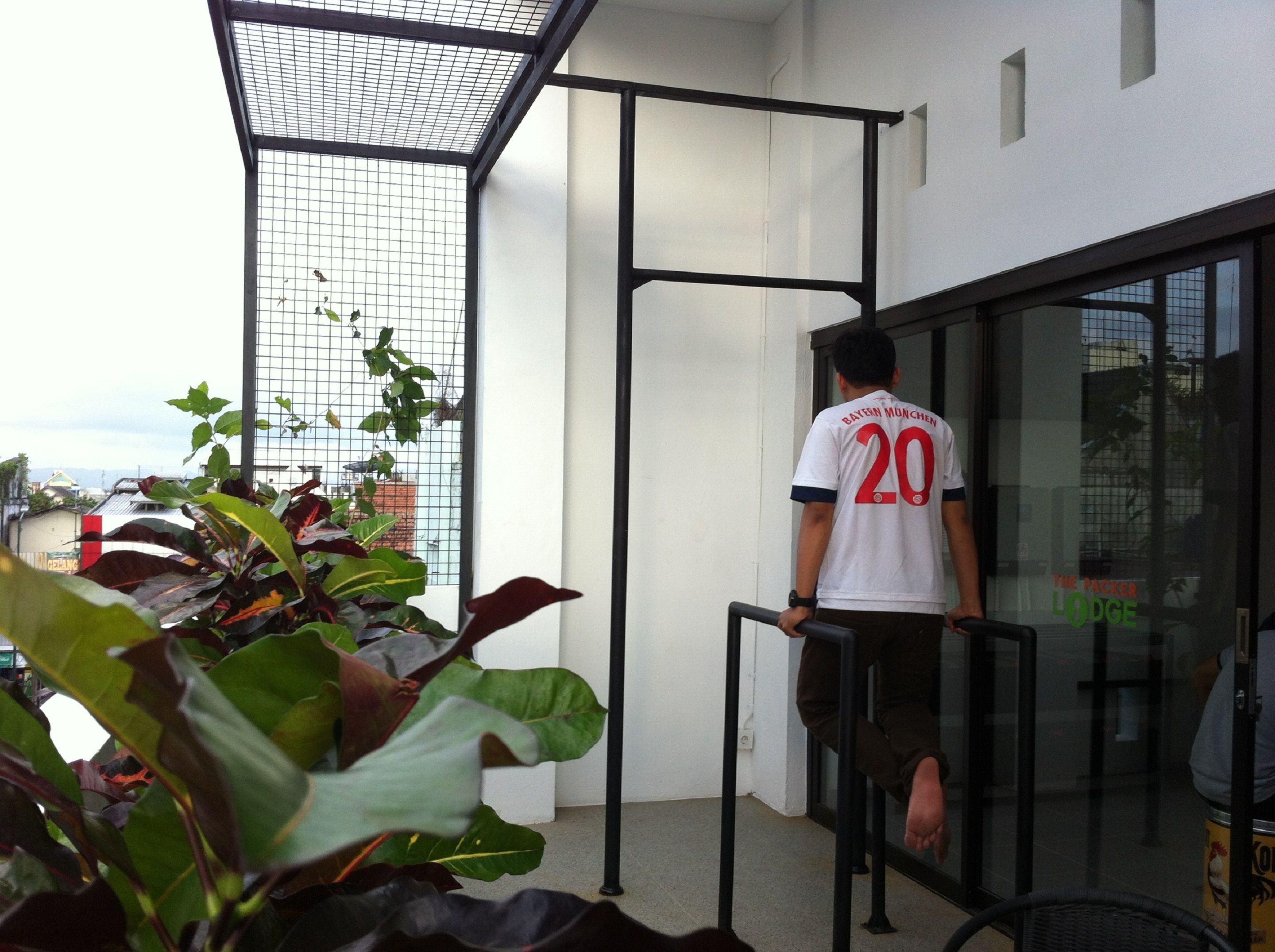 Una persona con camiseta deportiva blanca, con el número 20 y la inscripción "Bayern Munich" en la parte trasera, parada frente a una puerta de cristal.